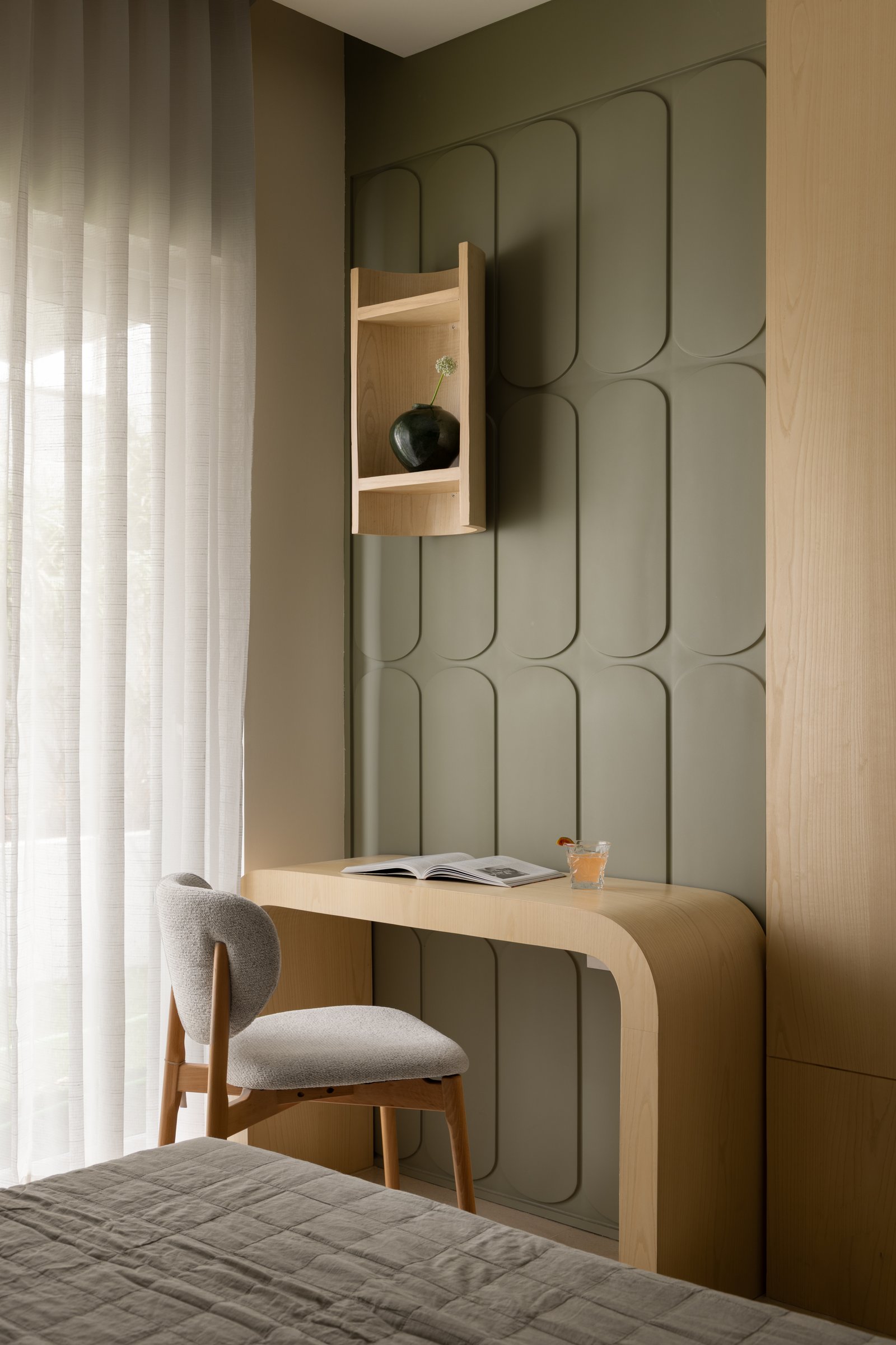 A sculpted sage-green wall panel defines the guest room study corner, its curved timber desk emerging directly from the surface