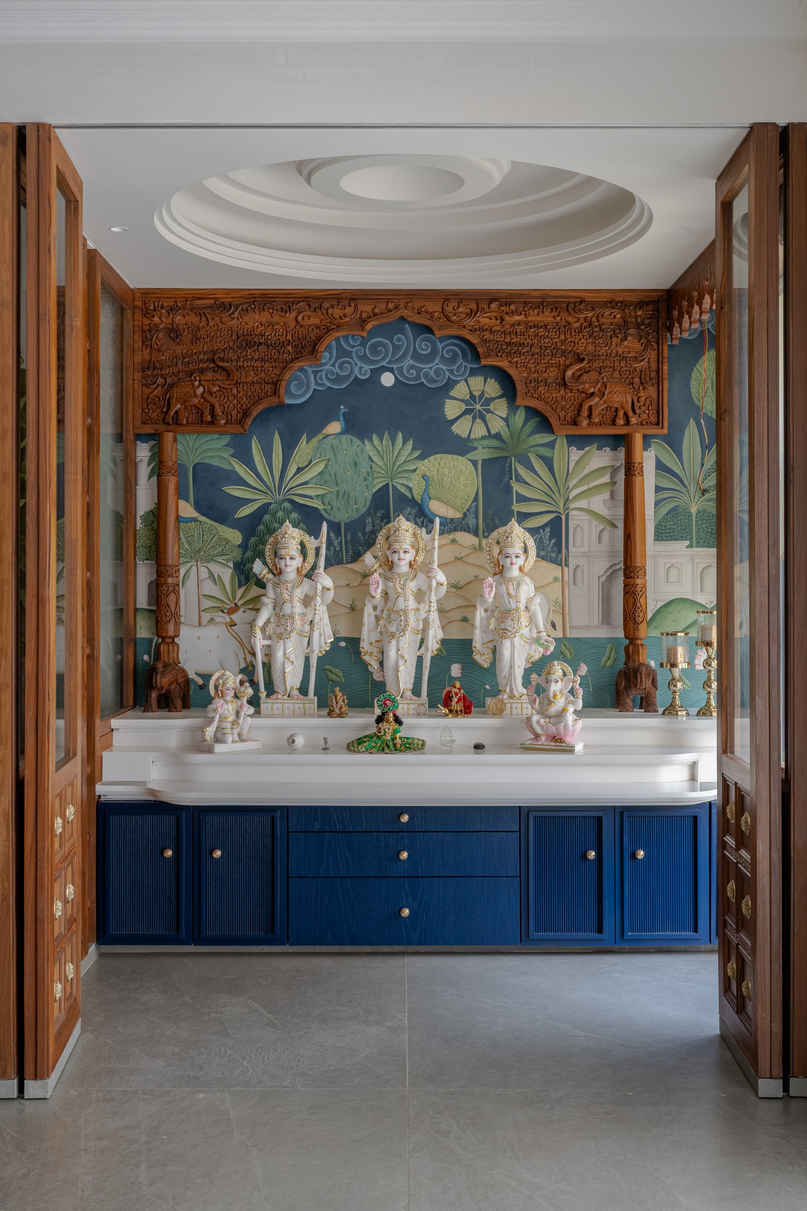 Wing B's pooja room, framed by a carved teak arch, with blue cabinetry below the Ram Darbar deities