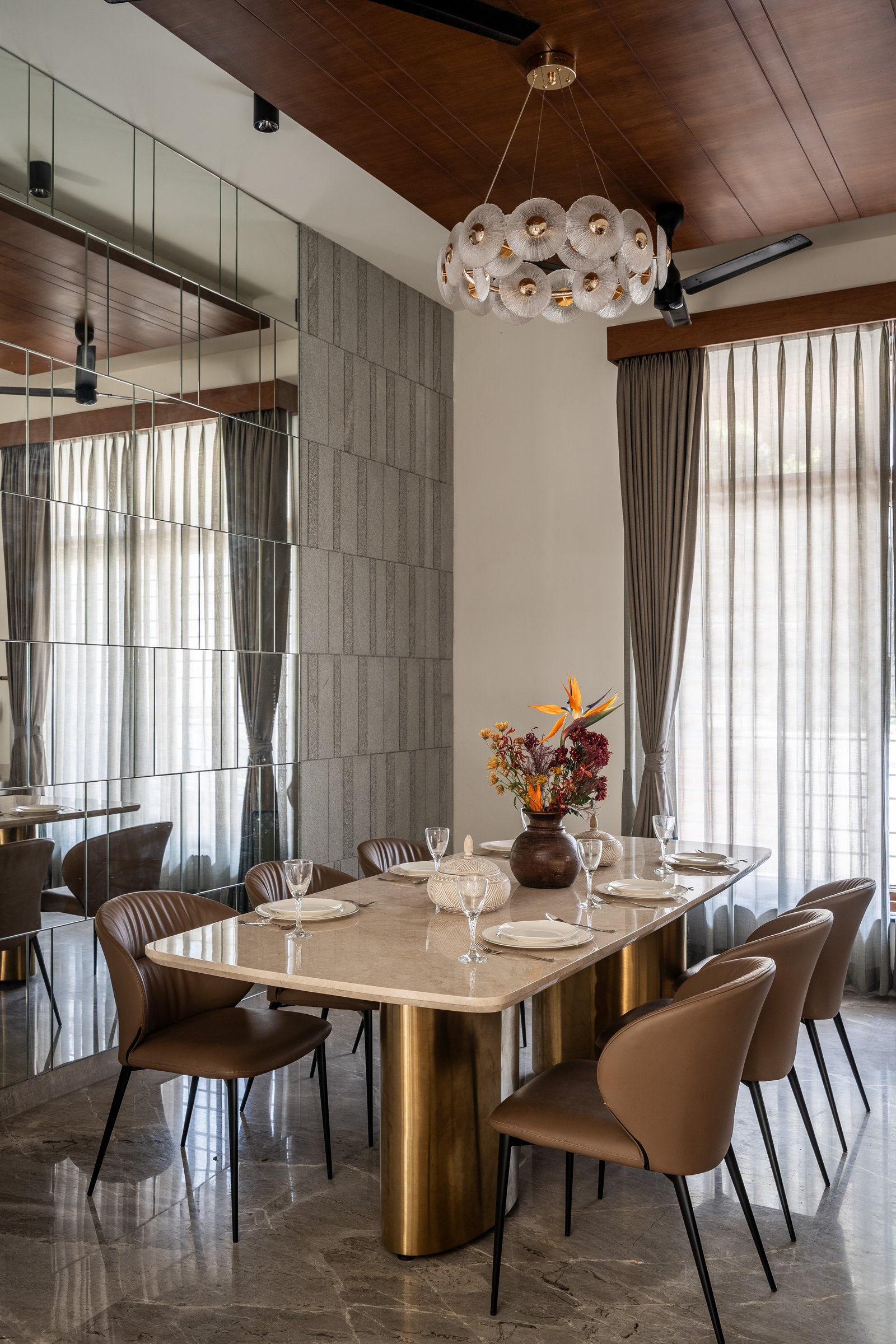 The dining room: a travertine table on a brass-clad pedestal, a mirrored wall, and a floral chandelier of pleated discs