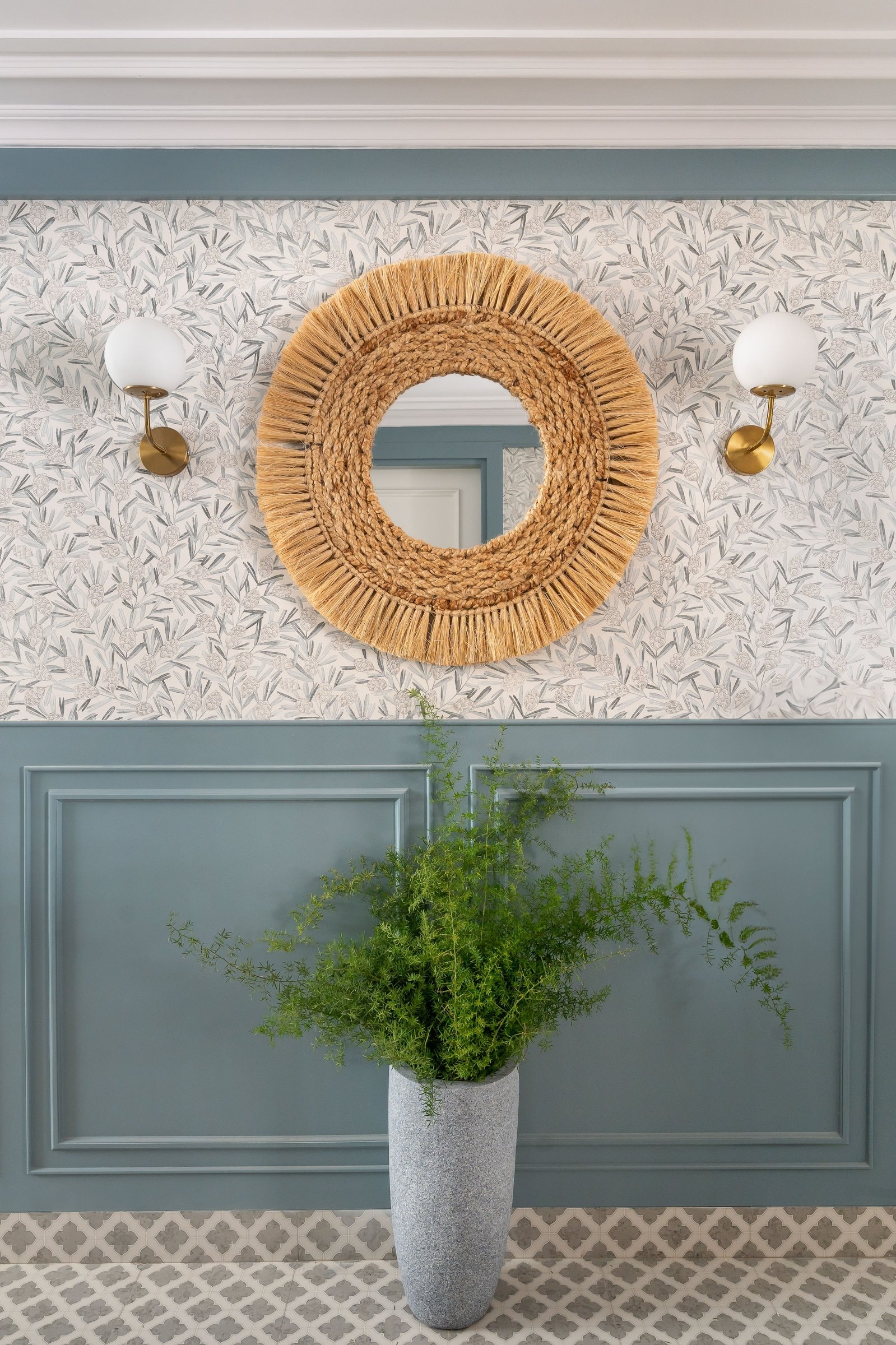A jute-fringed mirror between brass sconces, three textures resolved in one quiet wall