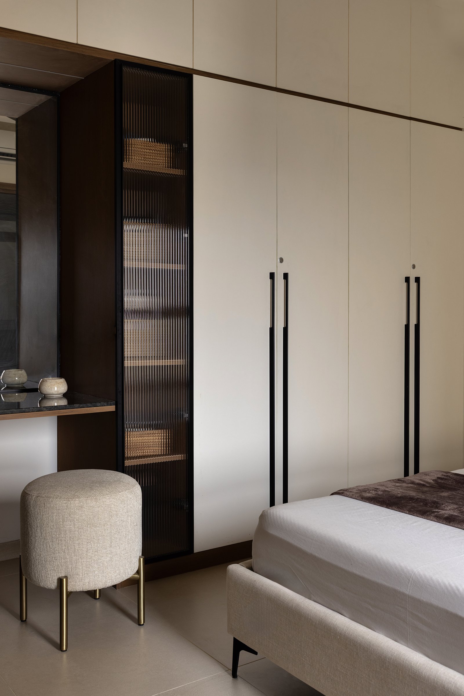 A wall of cream wardrobes with elongated black-and-brass pulls turns into a small, monastic dressing corner
