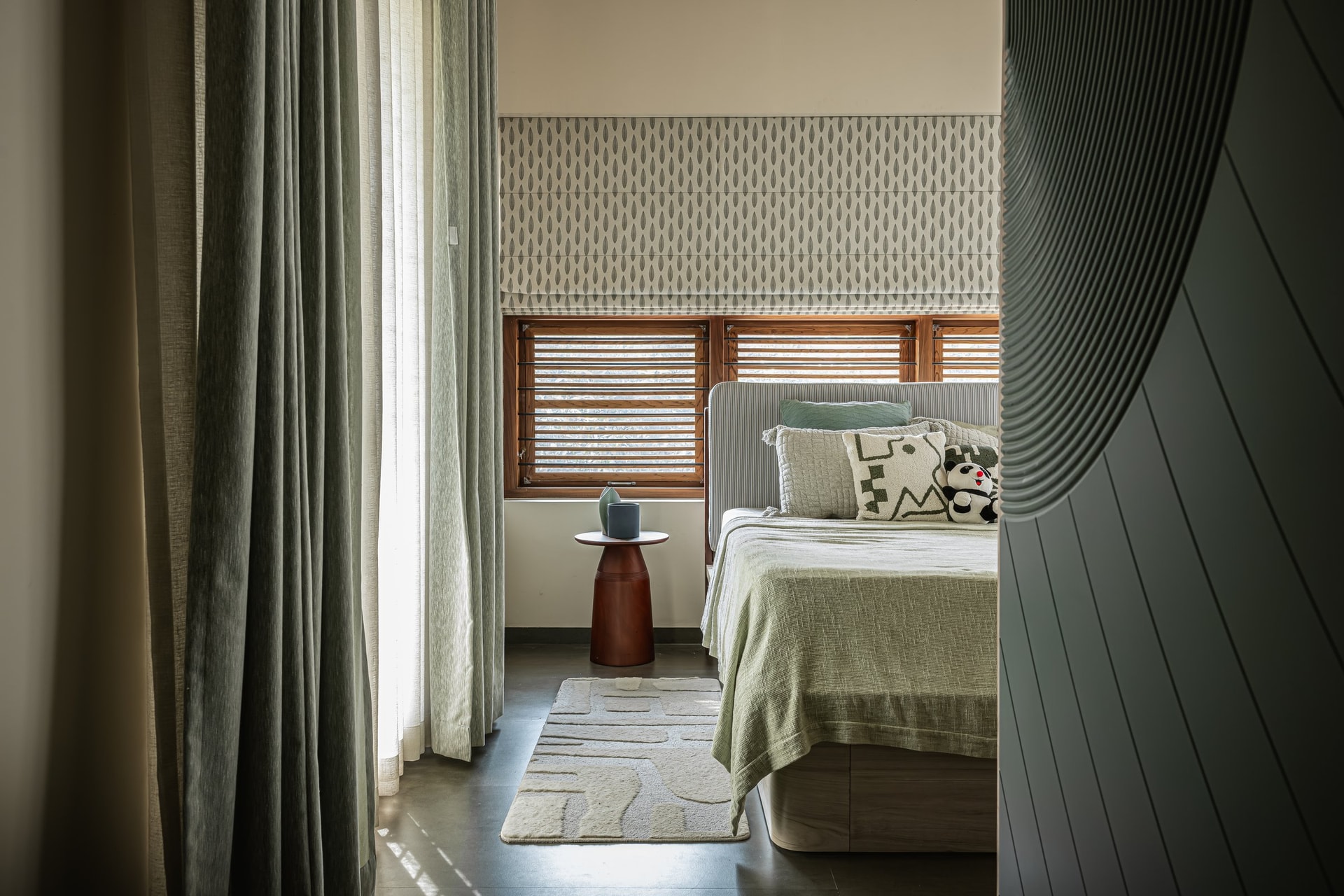 The master bedroom, where a patterned roman blind and layered sage textiles shift the home's register from communal to quiet