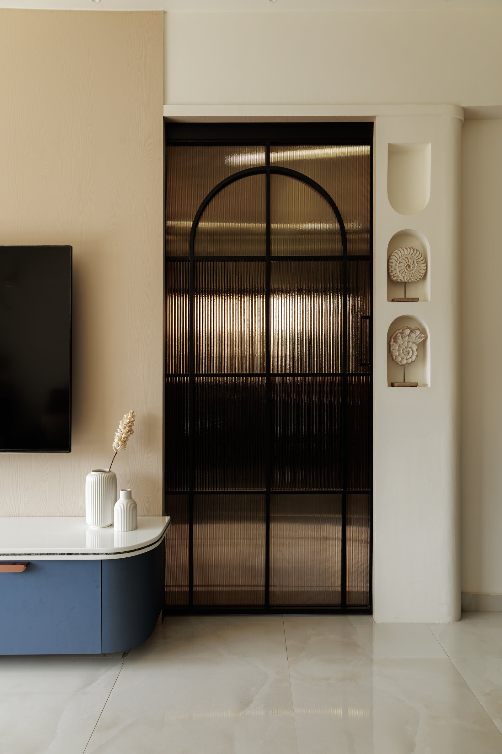 The threshold between living room and kitchen, framed by a reeded glass-and-metal sliding door and a pier of arched niches displaying ammonite-form sculptures