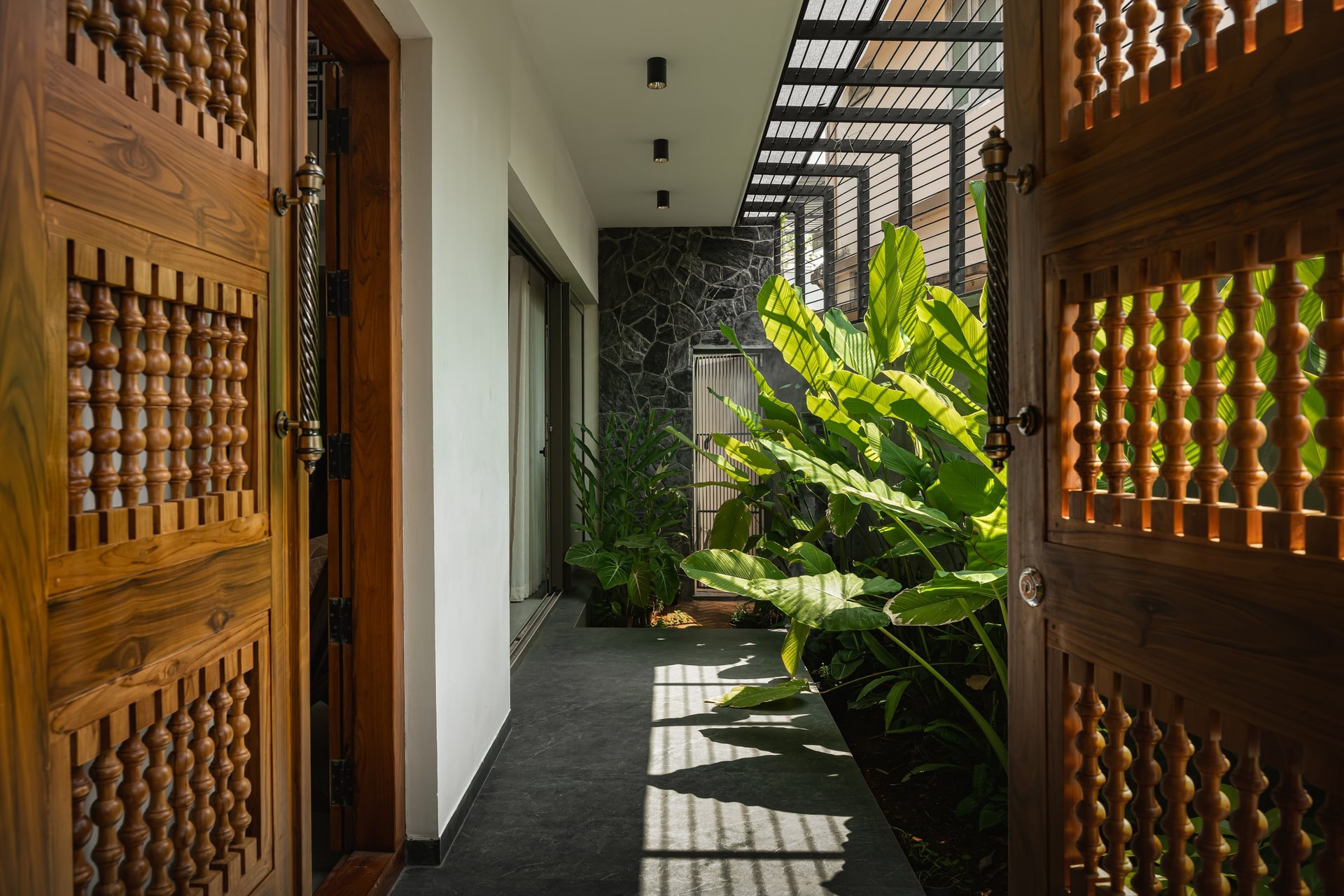 The entry passage: carved spindled doors, a planted strip, and a steel pergola translating Kerala light into striped shadow