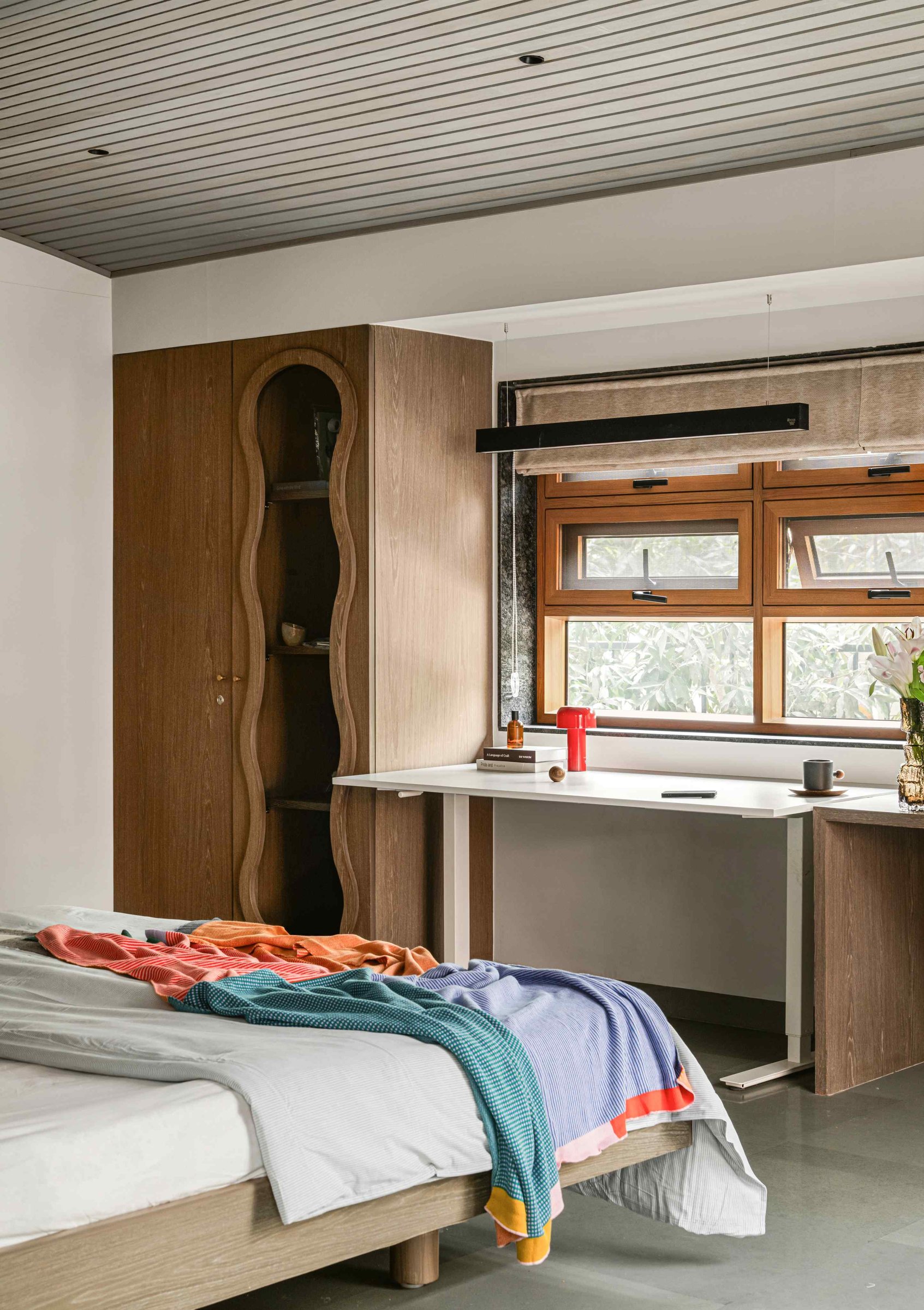 The same bedroom from the work corner: a wall-mounted desk, slim linear pendant, and a wave-carved wardrobe slot