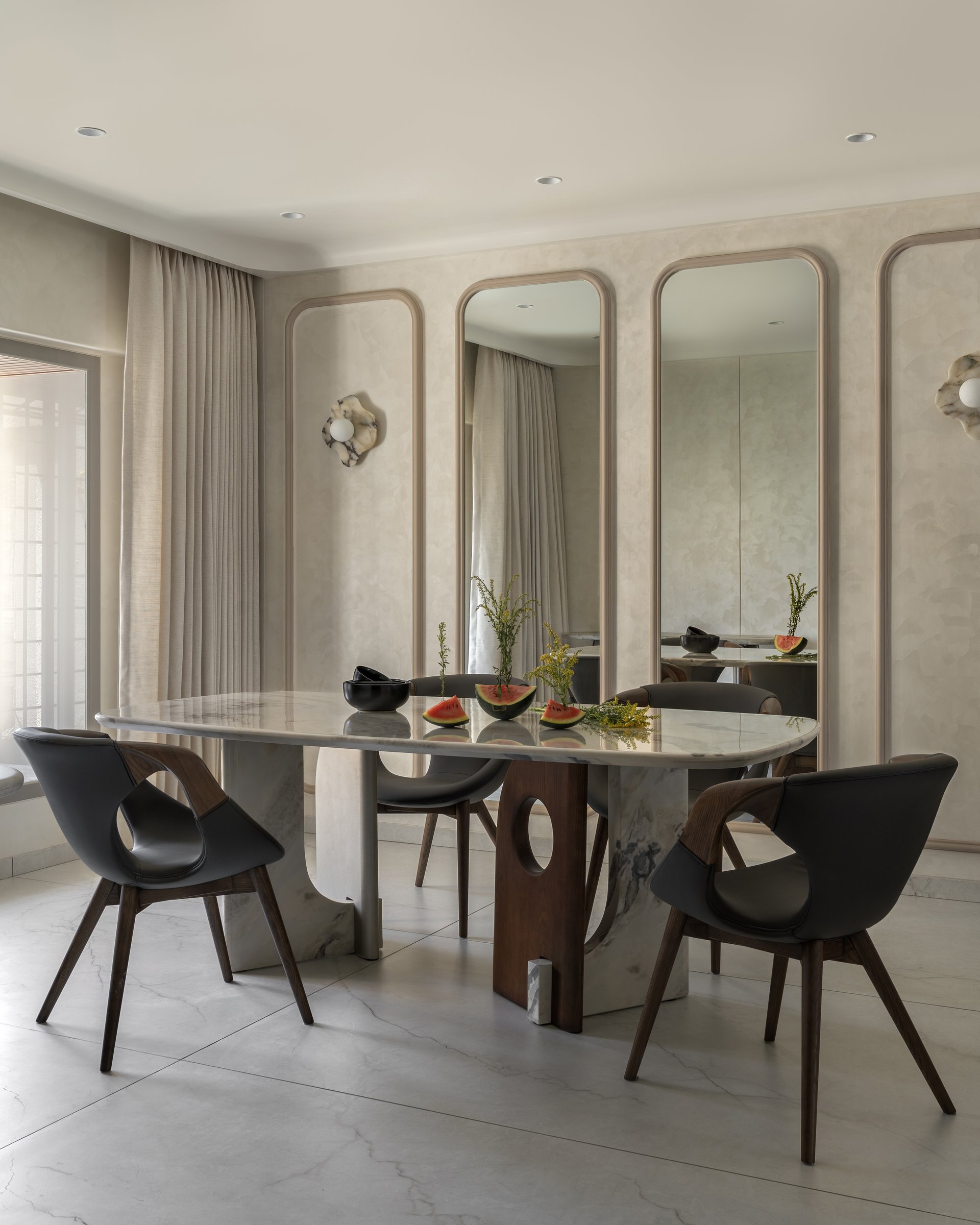 The dining room's wall of arched mirror panels in soft plaster mouldings, framing a stone table and walnut chairs
