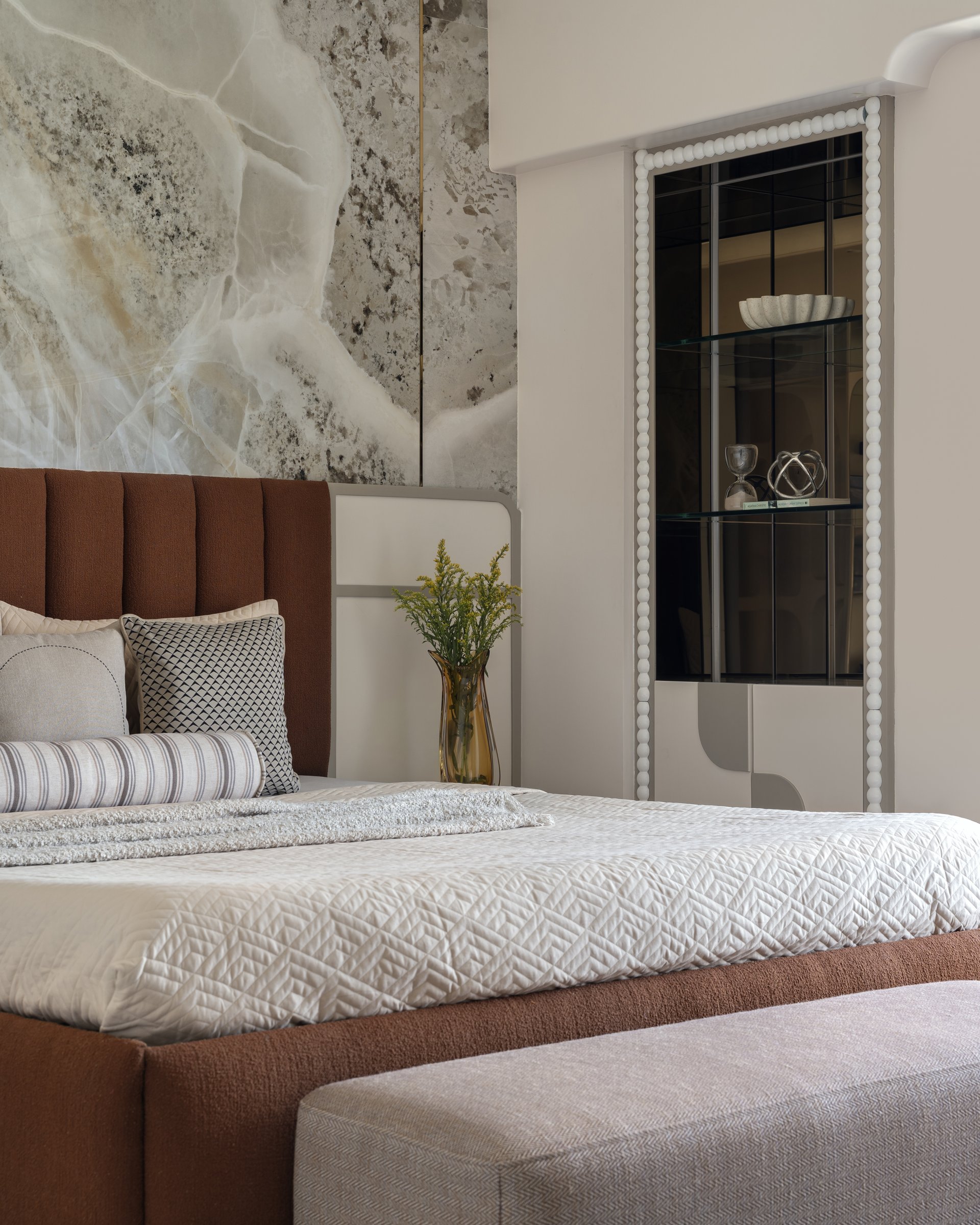The primary bedroom: a rust boucle headboard against figured stone cladding, with a beaded white vitrine alongside