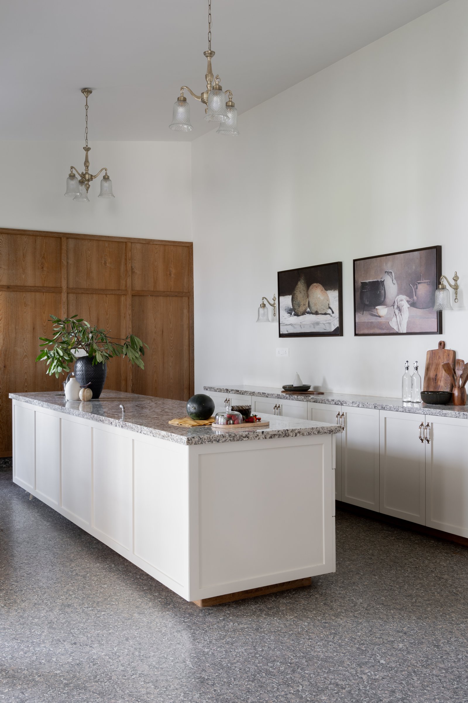 In the kitchen, two brass-and-glass chandeliers hang against a double-height white wall above the granite island
