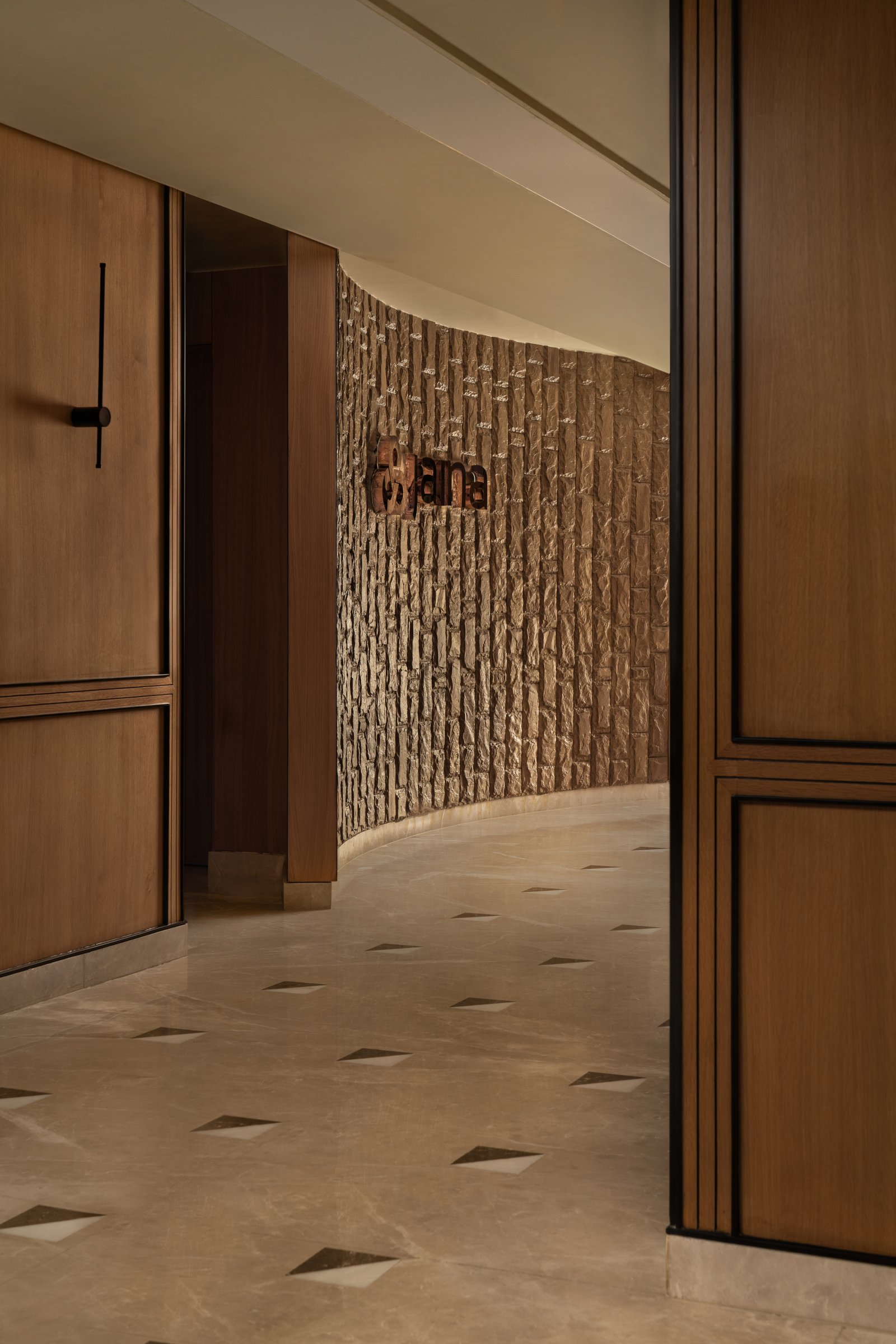 The curved stone wall behind reception, hand-chiselled and holding the Jaina insignia in raised bronze
