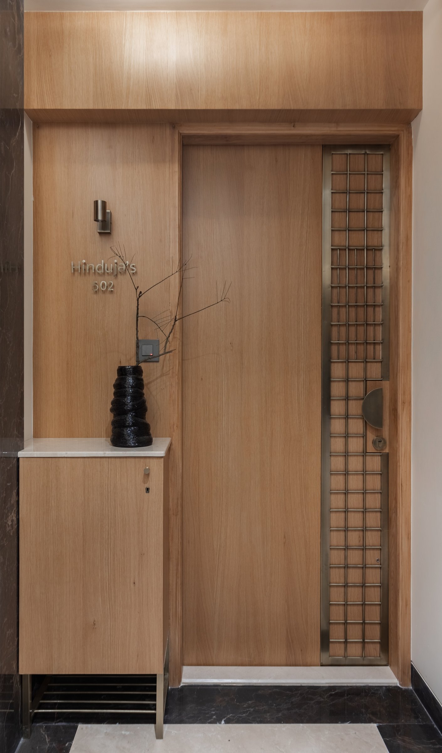 The entry door, where warm oak panelling and a tall brass grille establish the apartment's material grammar before the threshold is crossed