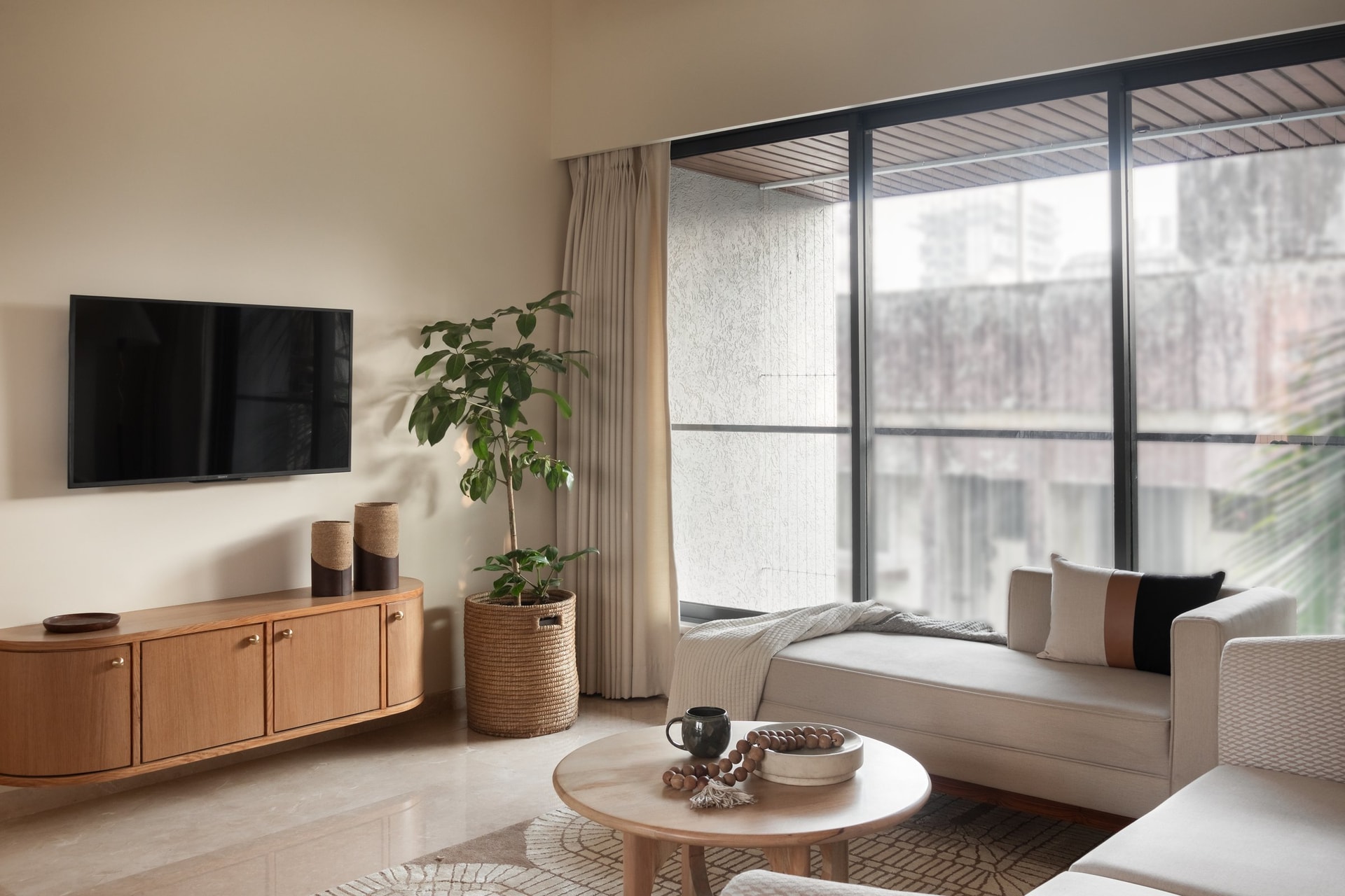 The living room, where a low cream daybed and a curved oak sideboard hold the apartment's tonal key beneath a generous picture window