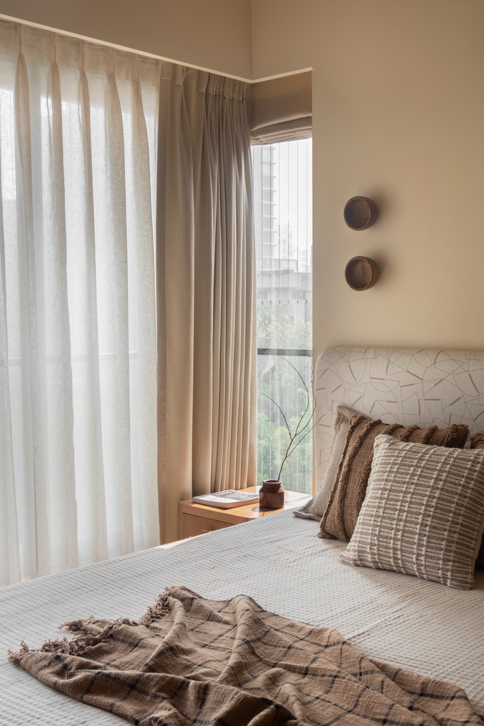To one side of the master bed, sheer linen curtains and small turned-wood sconces let the green of the neighbourhood enter without diluting the room's warmth
