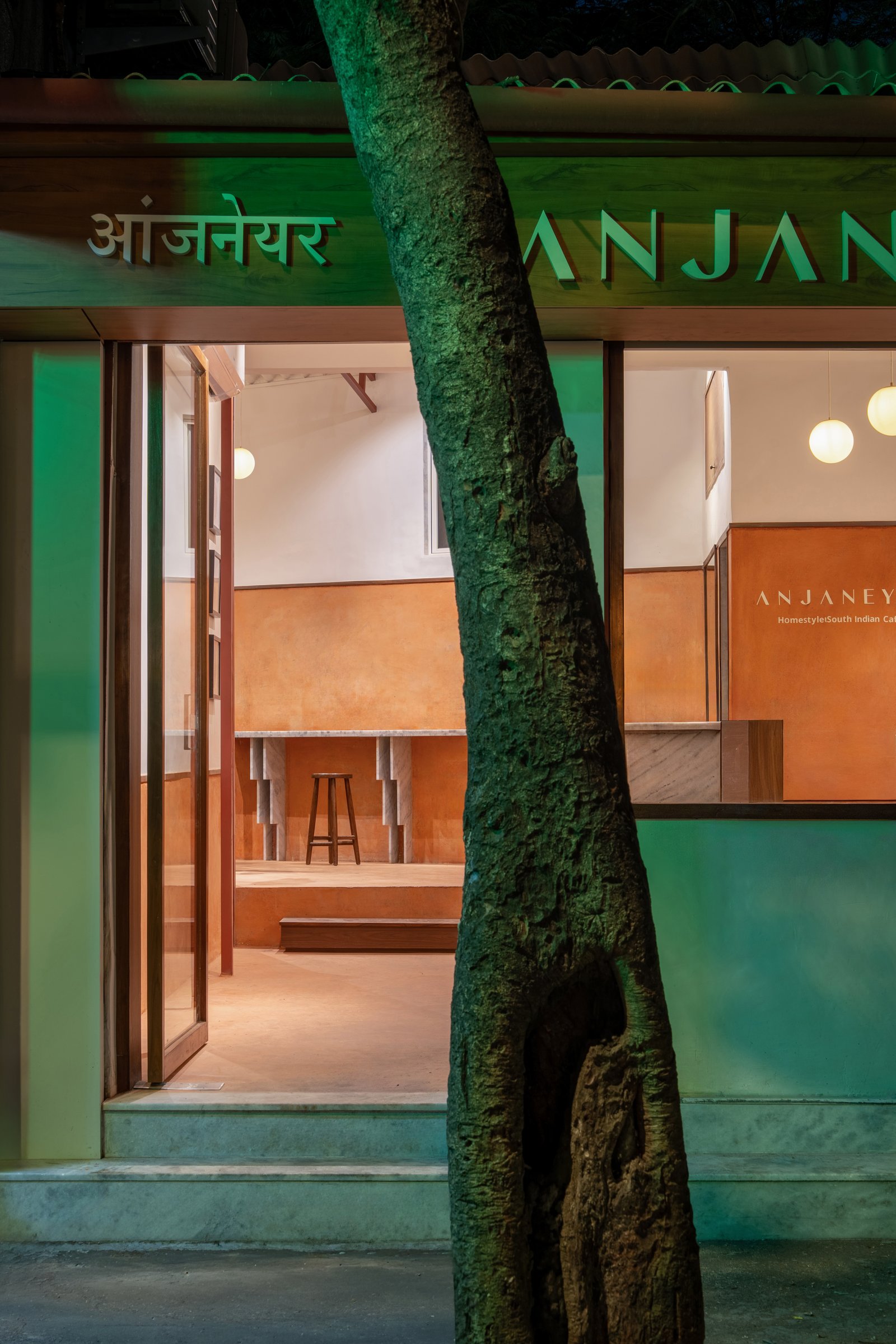 The façade at dusk, the existing tree retained at the threshold and the green signage marking the entry