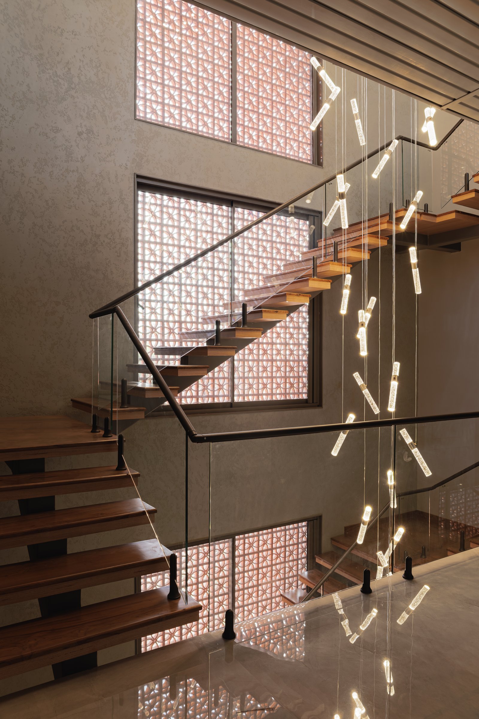 From above, a cascade of bar pendants drops through the double-height void against the luminous brick jaali screen