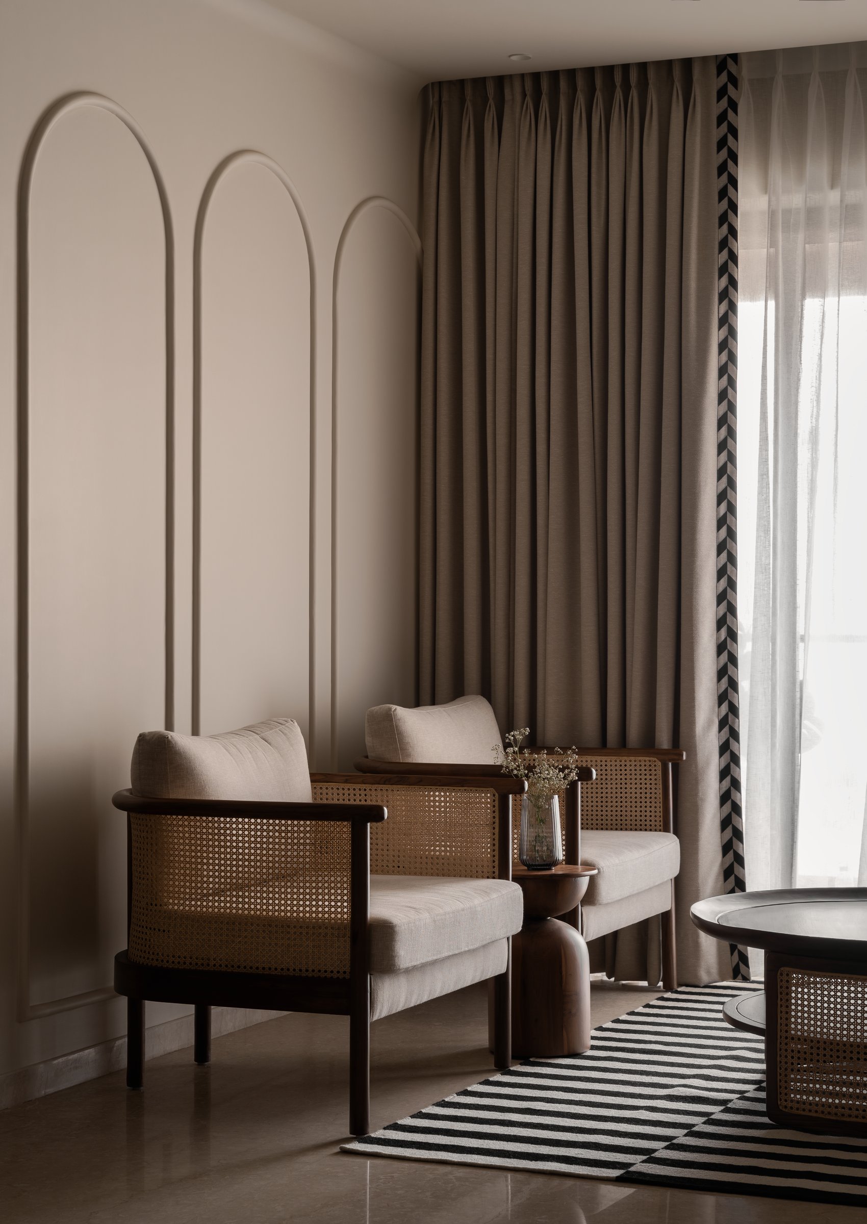 The living room, where slim arched wall mouldings and cane-and-wood armchairs hold a calibrated conversation with chevron-trimmed sheers