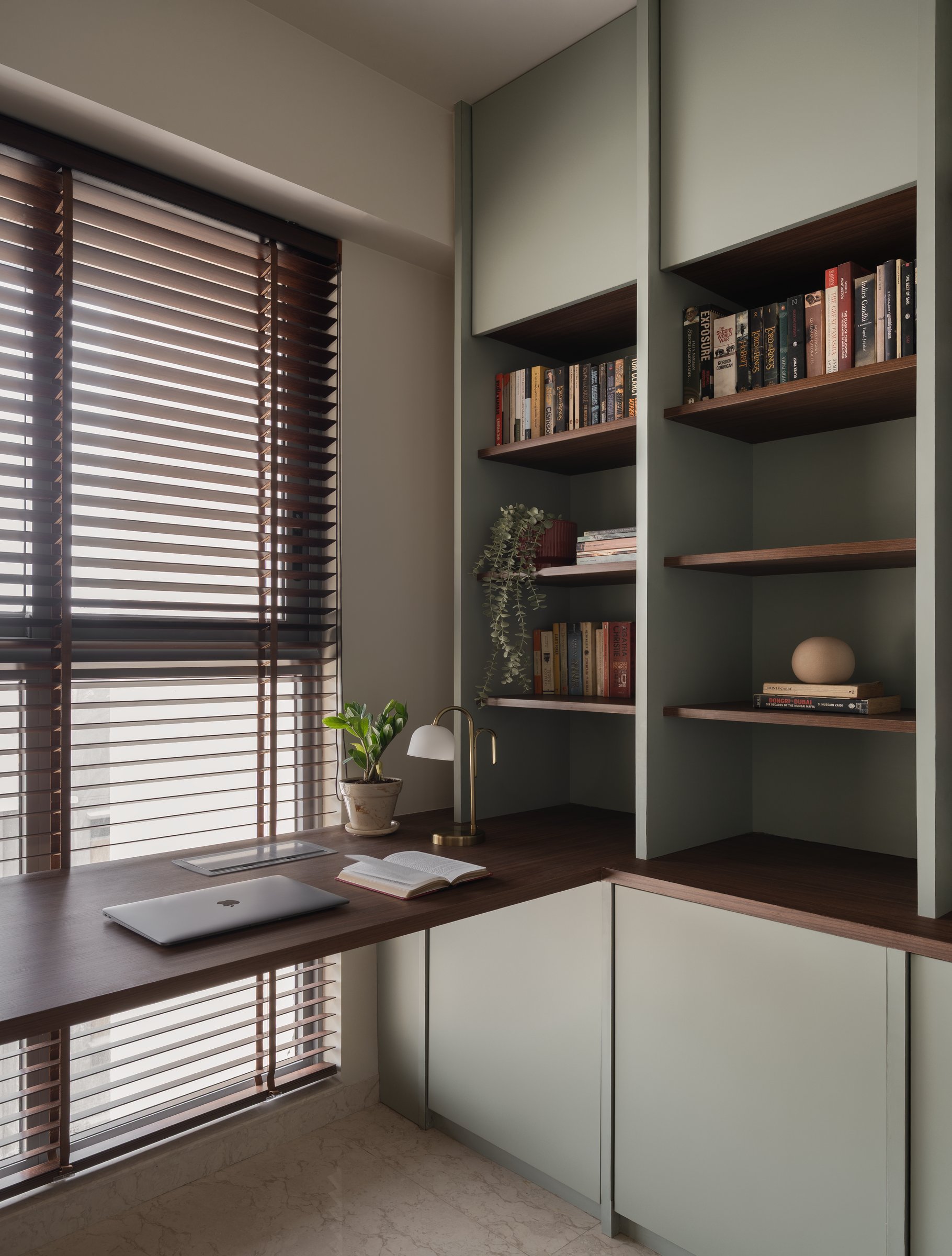 The home's study, where walnut joinery meets sage cabinetry and timbered Venetian blinds throw striped light across the floor