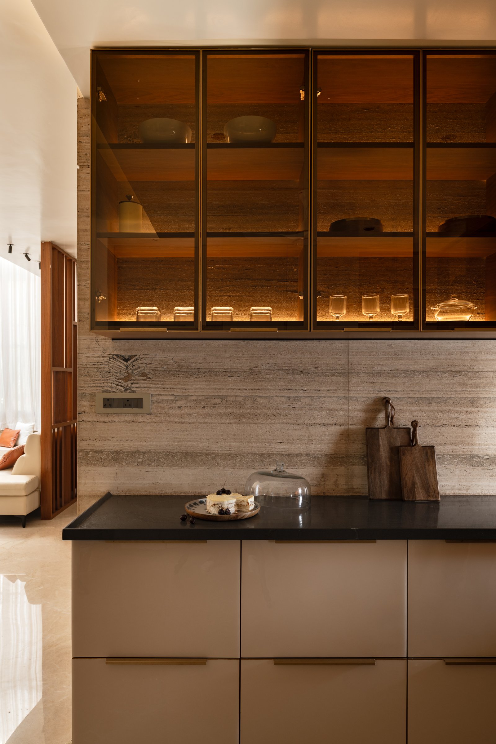 The kitchen: brass-framed glass uppers glow against a travertine backsplash, with soft taupe cabinetry below