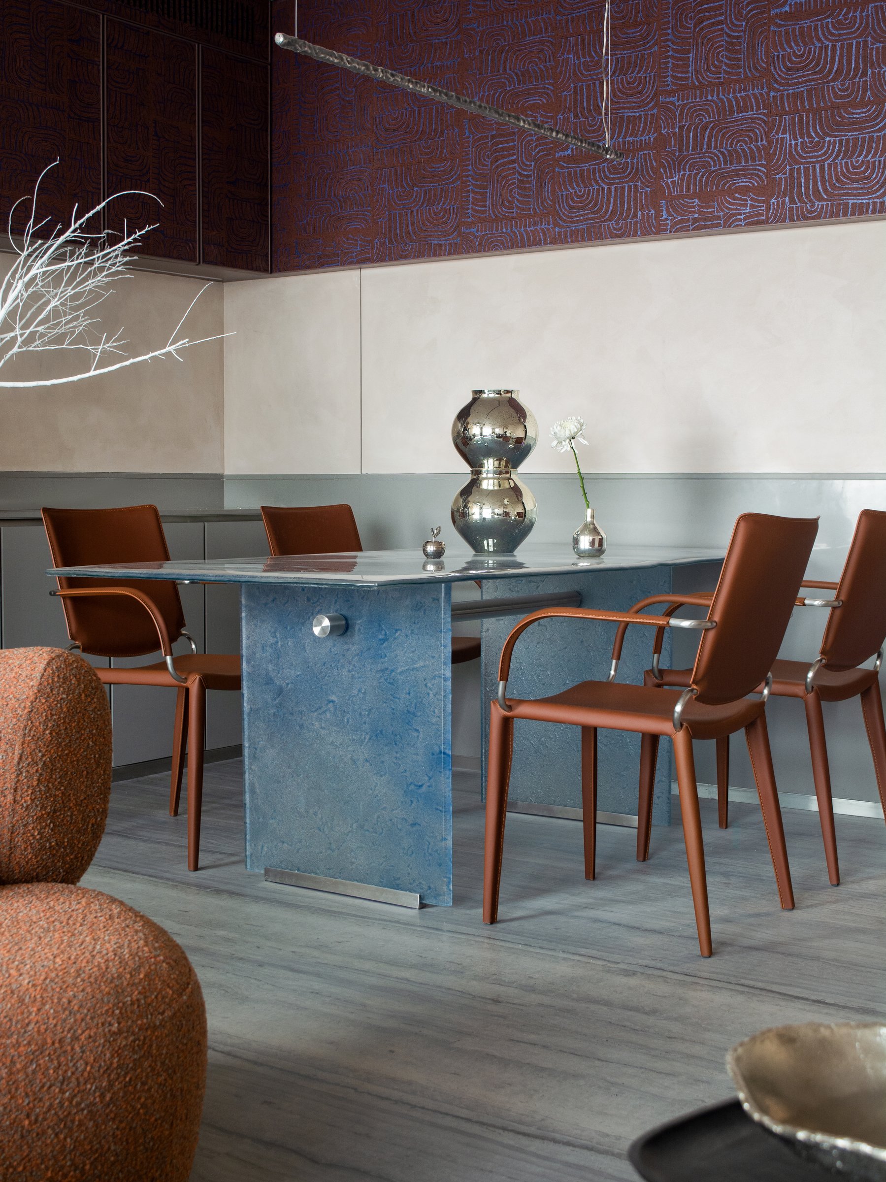 The dining area, anchored by a poured-glass table in pale blue and tan leather chairs, beneath a patterned panelled upper wall