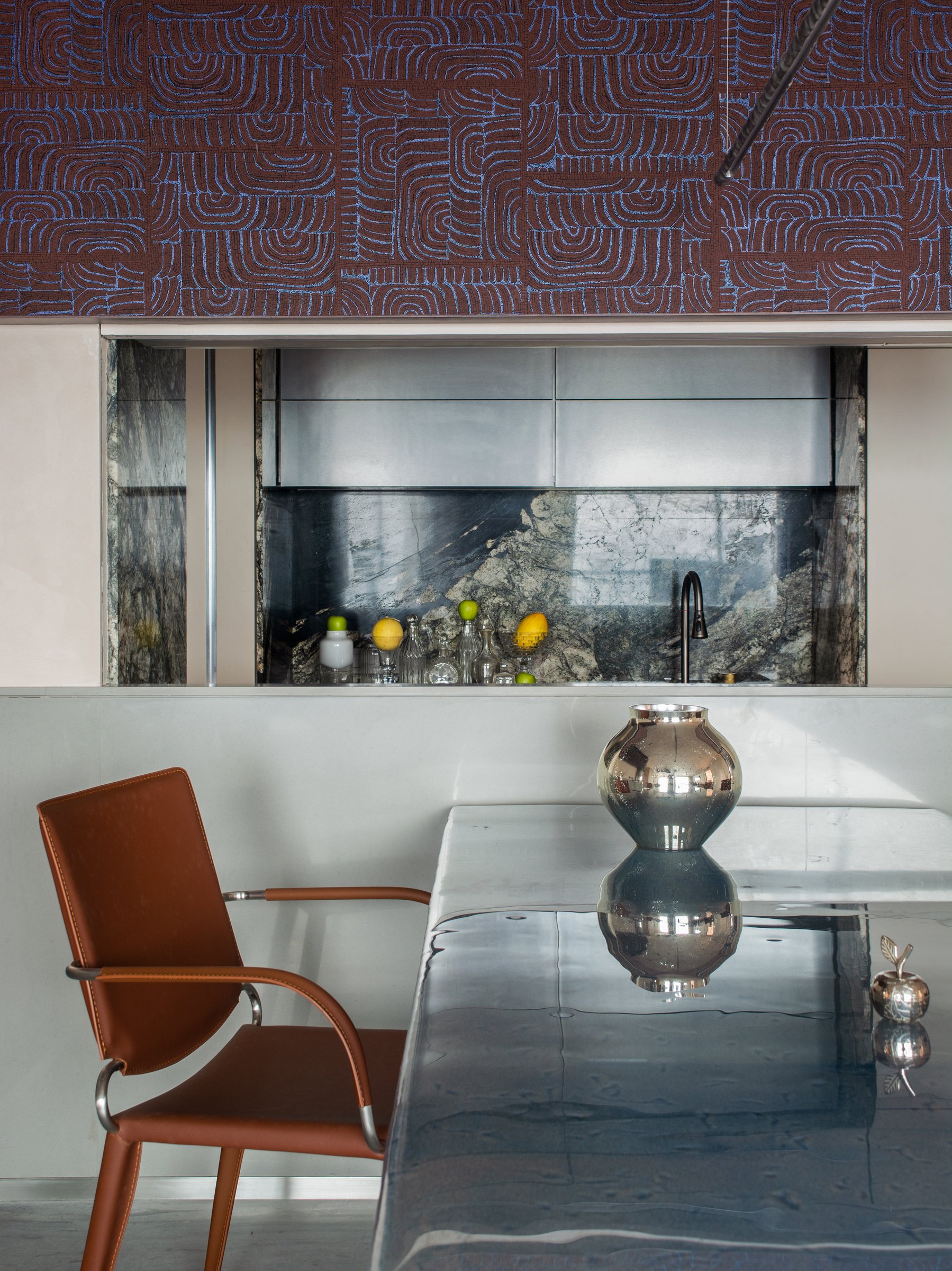 A detail of the glass tabletop, with the kitchen pass-through and veined dark stone backsplash visible beyond