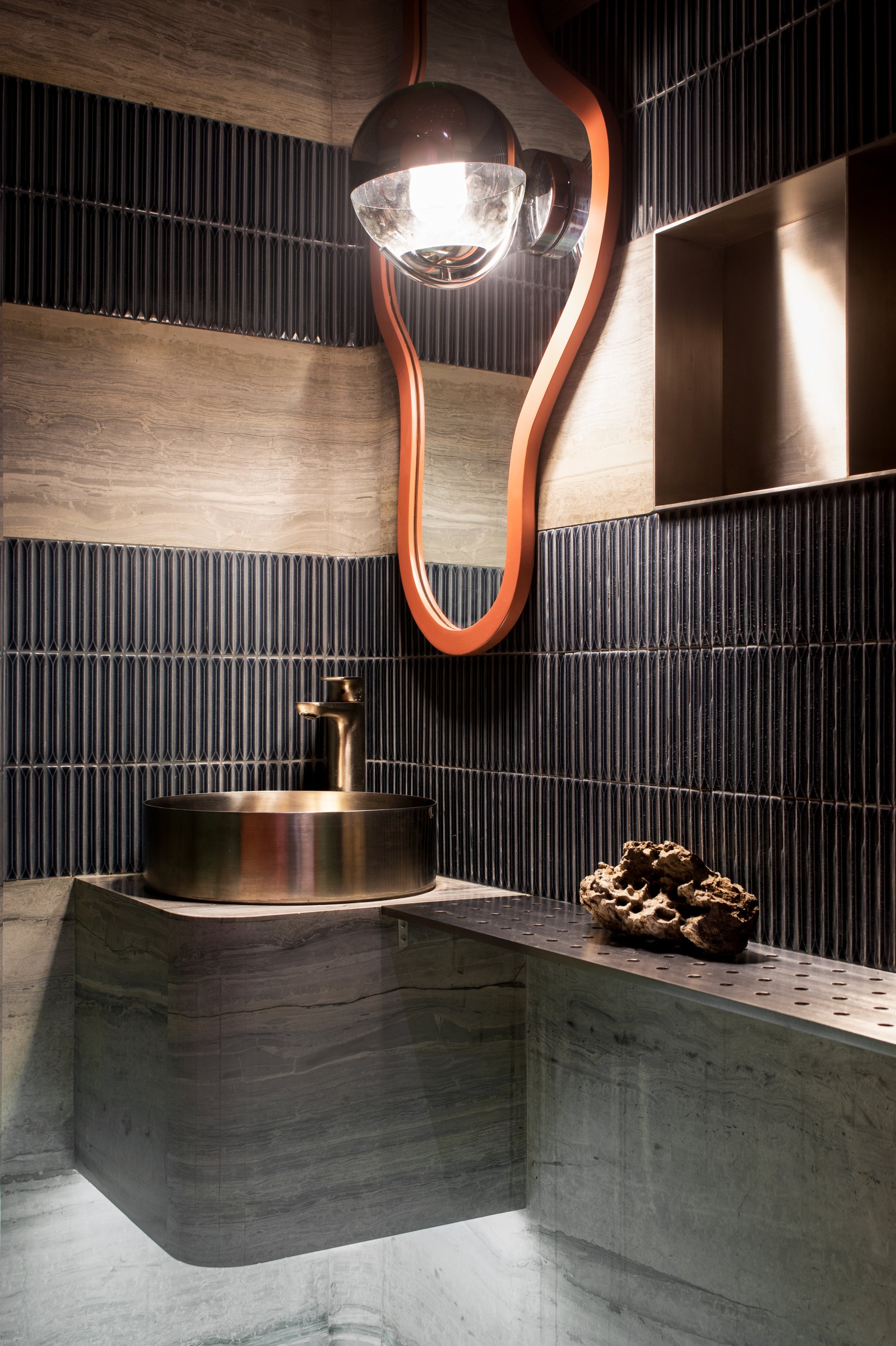 The powder room, where ribbed dark tiles meet a wavy orange-framed mirror and a brass vessel sink