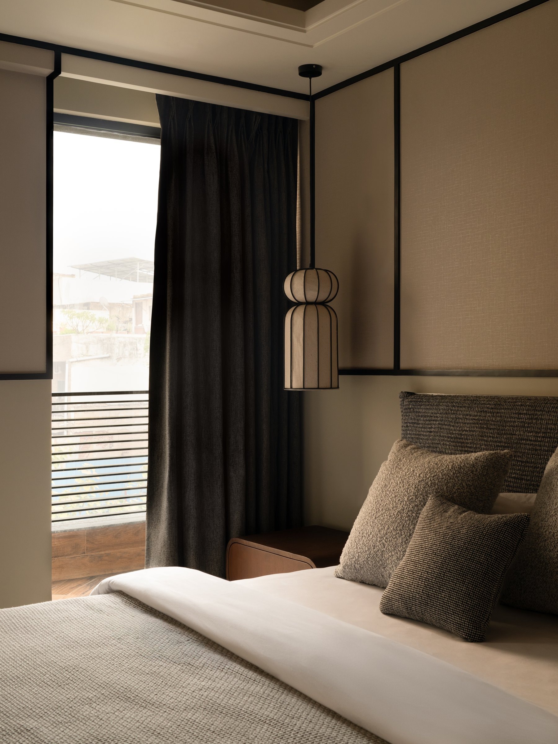 The bedroom opens to a balcony through floor-to-ceiling glazing dressed in pleated dark drapery, daylight entering as a soft column along one edge