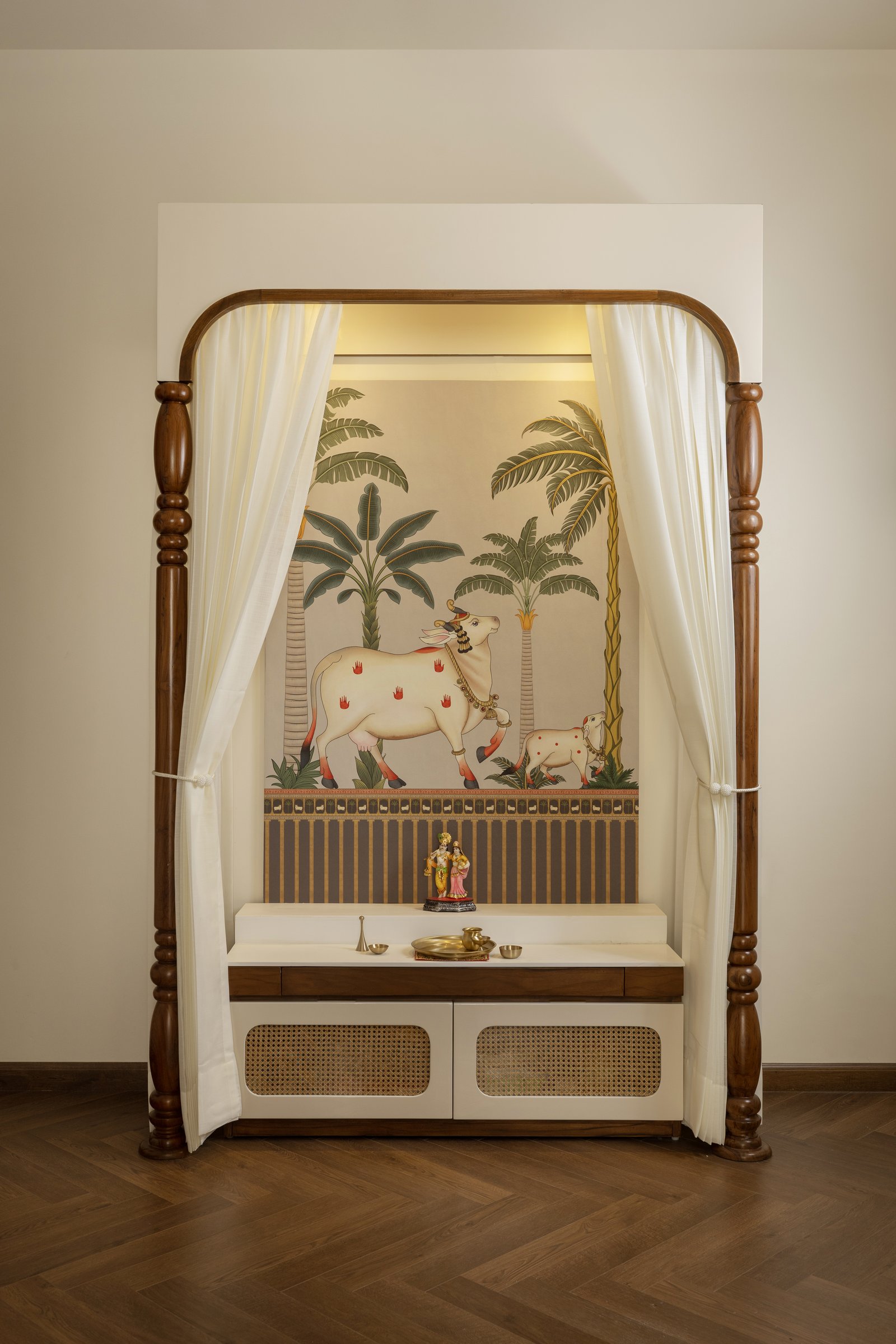 The pooja niche, framed by turned wooden columns and white sheers, with a Pichwai-inspired mural behind a cane-fronted counter