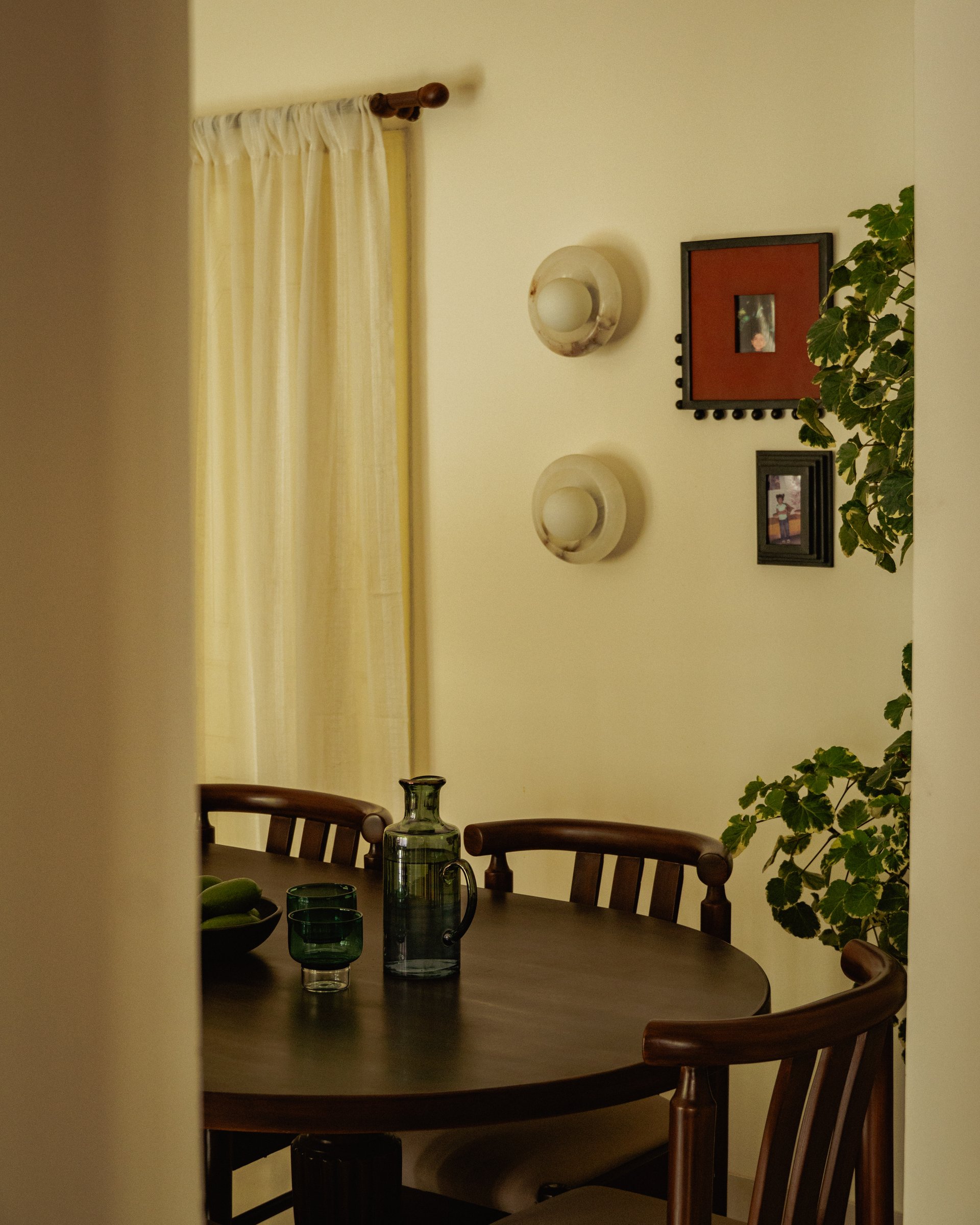 Framed through the doorway, the dining space reads closer to a parlour than a formal room, with sheer curtains and small framed portraits