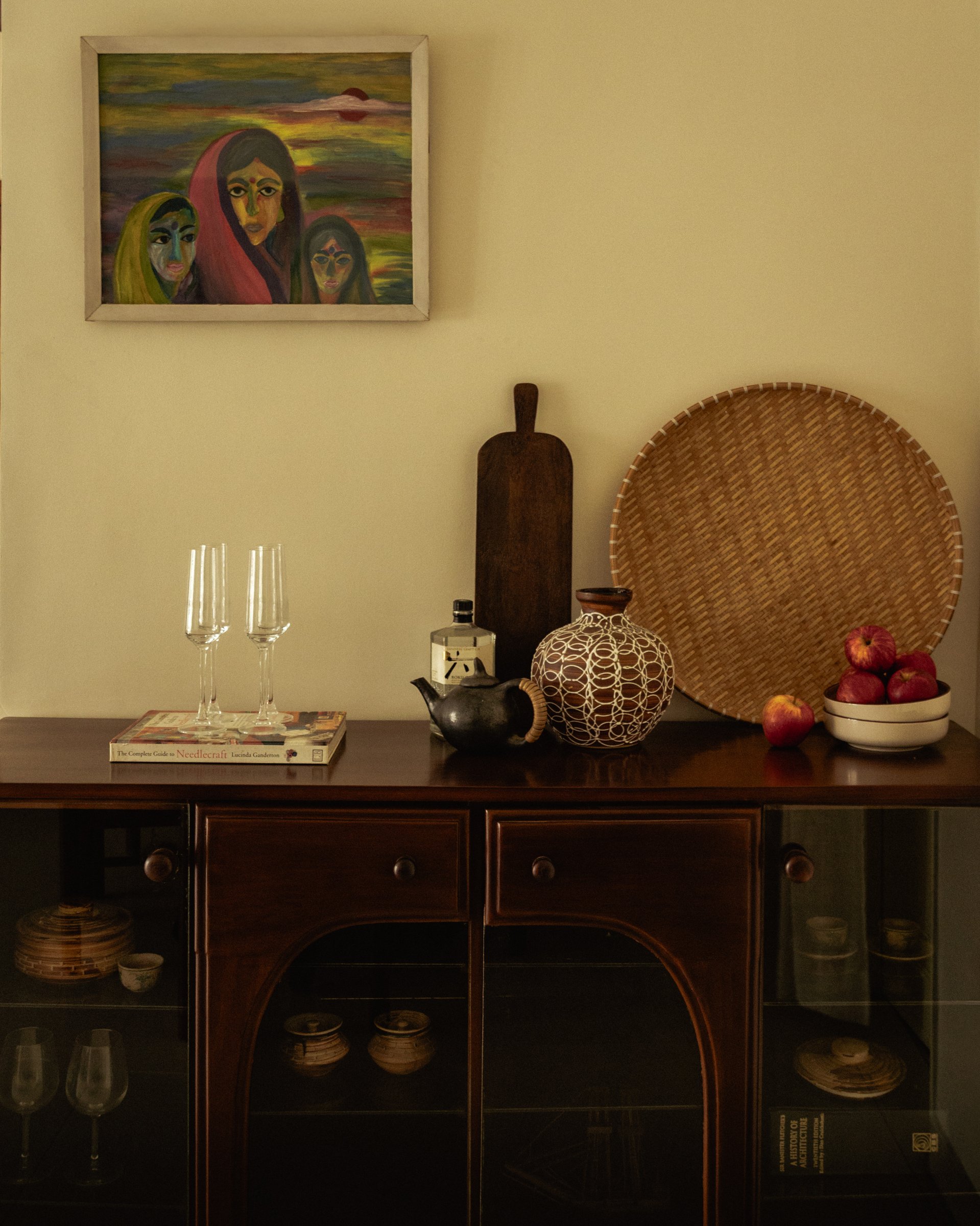 The sideboard beside the dining table, where a woven bamboo tray, a patterned round vessel, and a painted canvas of three figures sit together