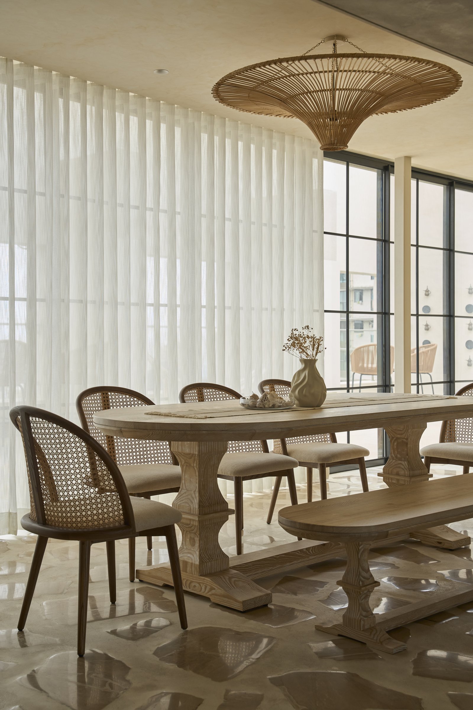 The dining area, where a pale timber trestle table and cane-back chairs sit beneath a wide woven rattan pendant