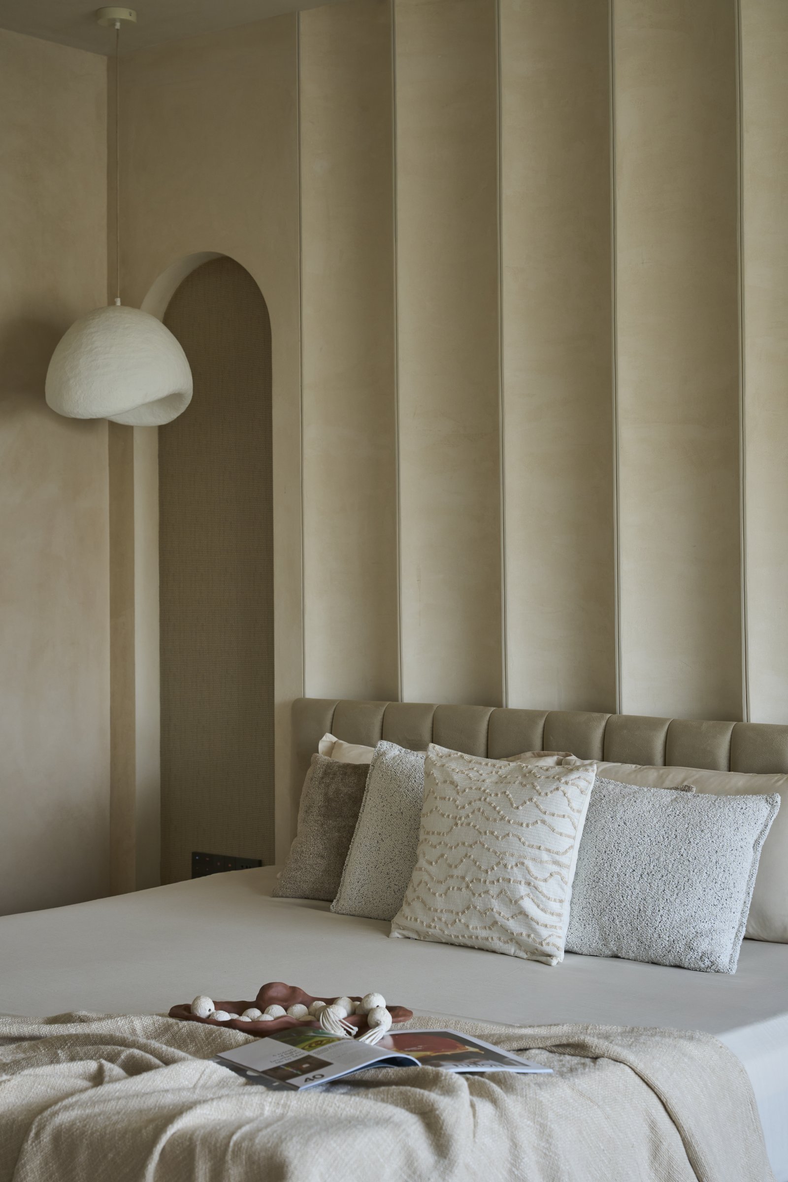 The bed wall, where a channelled headboard rises the full height beside an arched niche and sculptural pendant