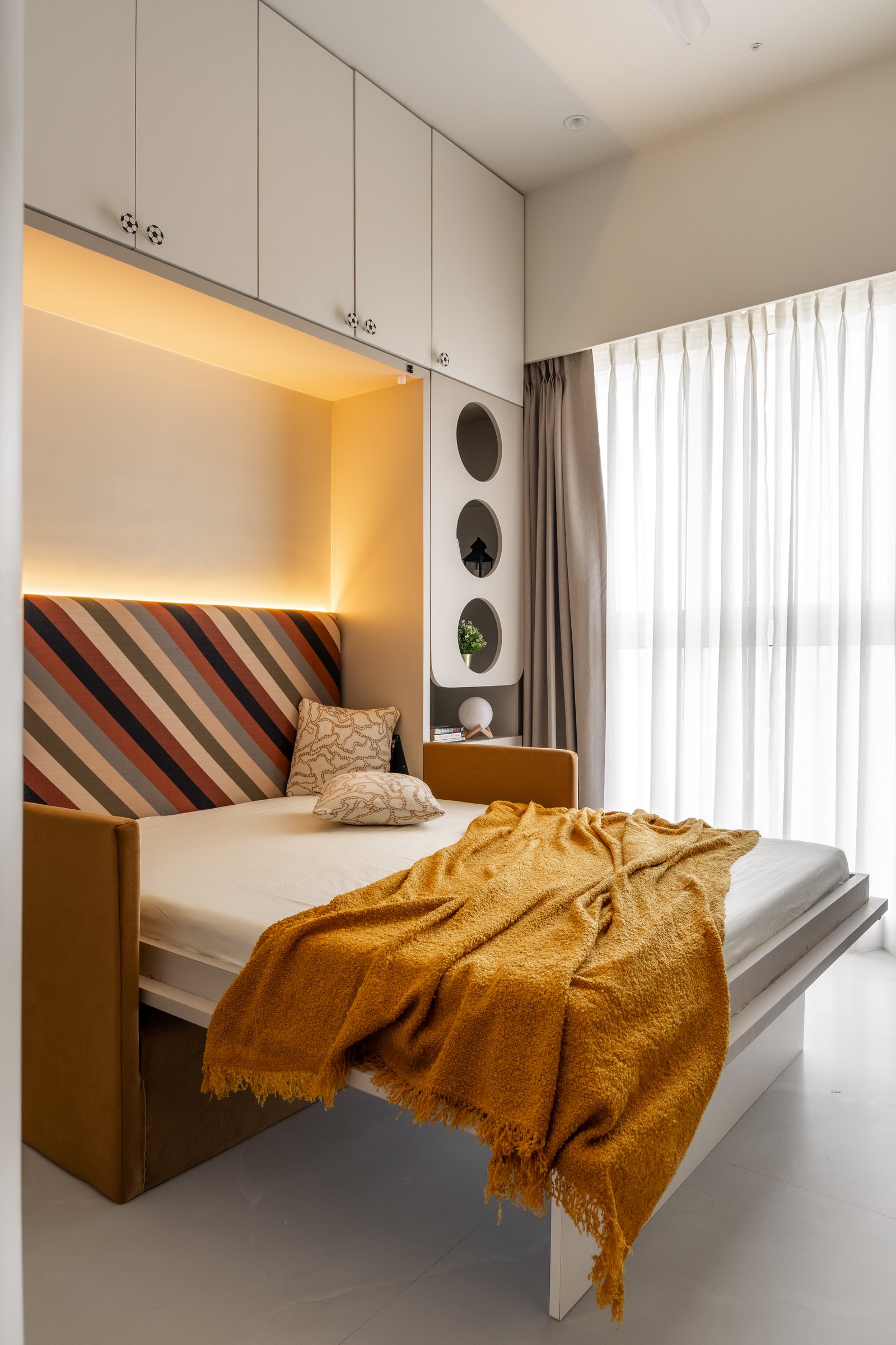 The son's bedroom, where a foldaway bed with a striped headboard tucks into a cove-lit niche, freeing the floor for daytime use