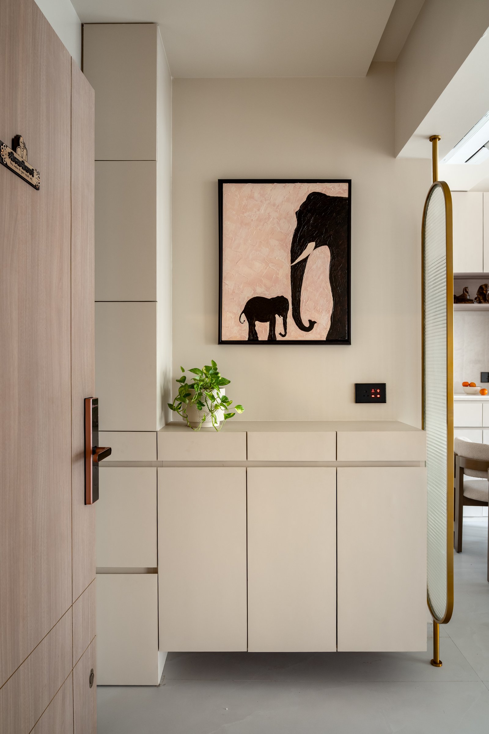The entry, where a wood-grain door opens onto a concealed storage wall, an elephant artwork, and a brass-framed screen filtering the view to the dining beyond