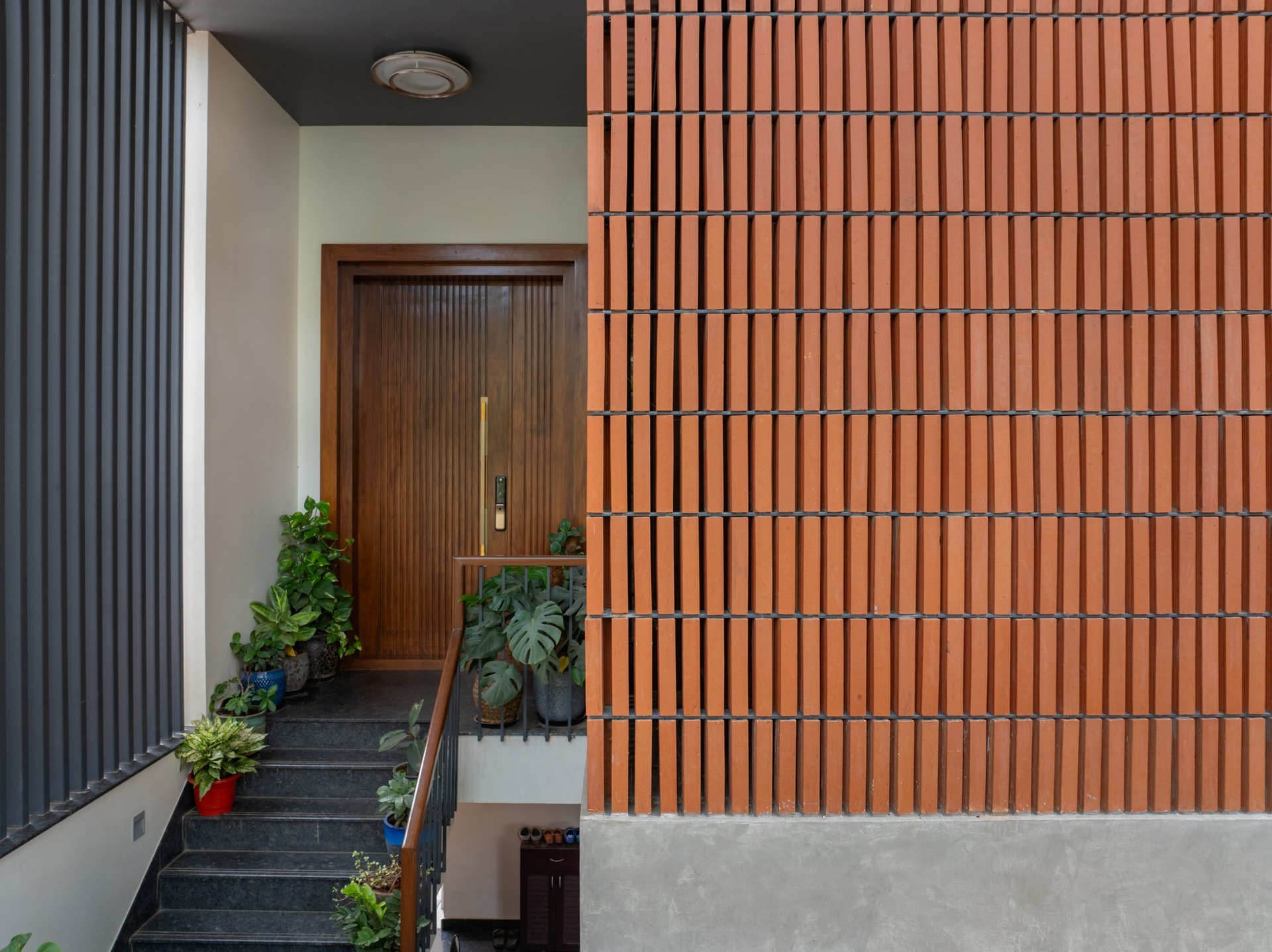 The entry threshold, where a perforated terracotta brick wall meets a slim aluminium fin screen along the granite ascent to the front door