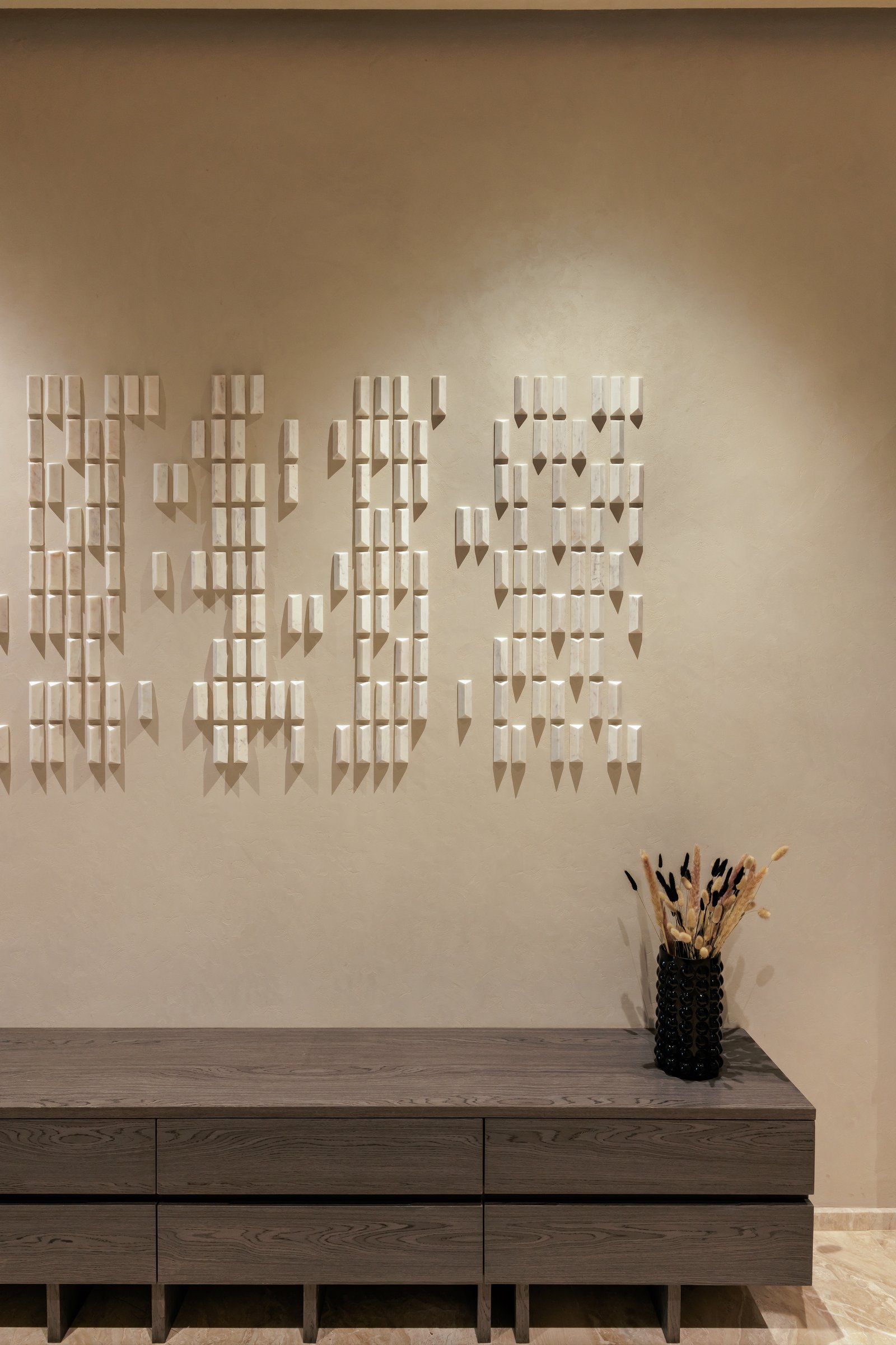 The foyer, where a low oak console anchors a wall of pale stone modules arranged like partial script across textured plaster