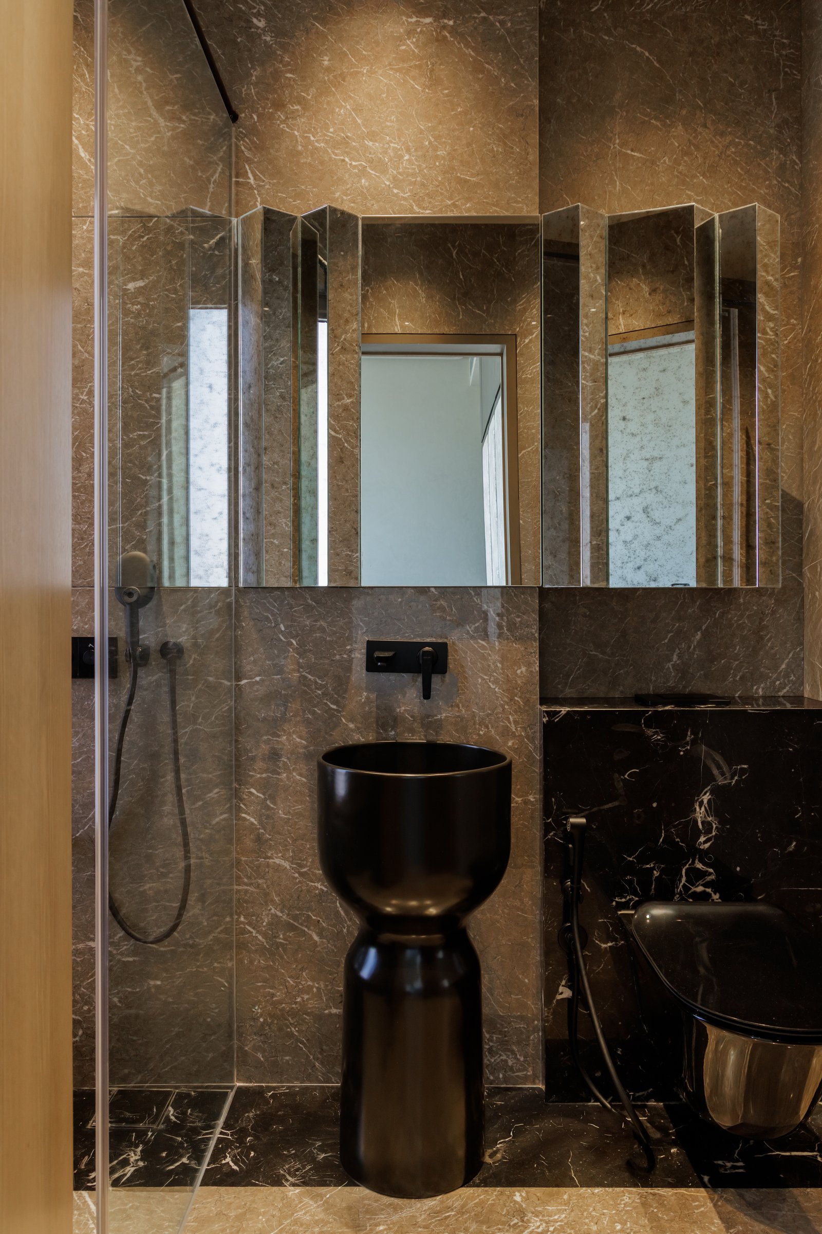A second bathroom, where warm-toned marble and a folded arrangement of antique-finish mirrors frame a dark pedestal basin