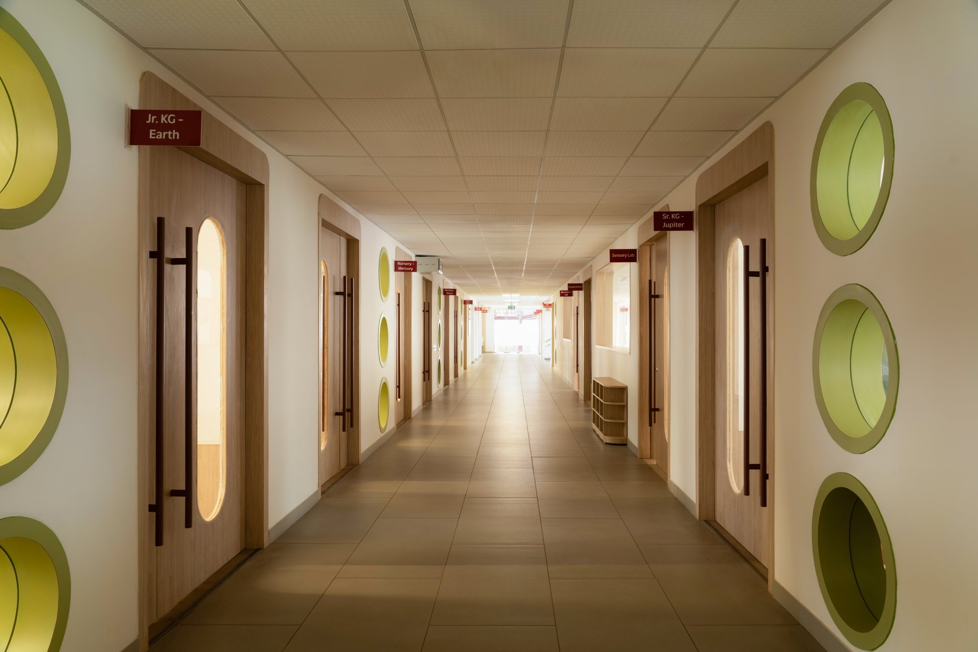 The primary corridor, lined with capsule-glazed oak classroom doors, green-ringed circular apertures and deep red signage tabs