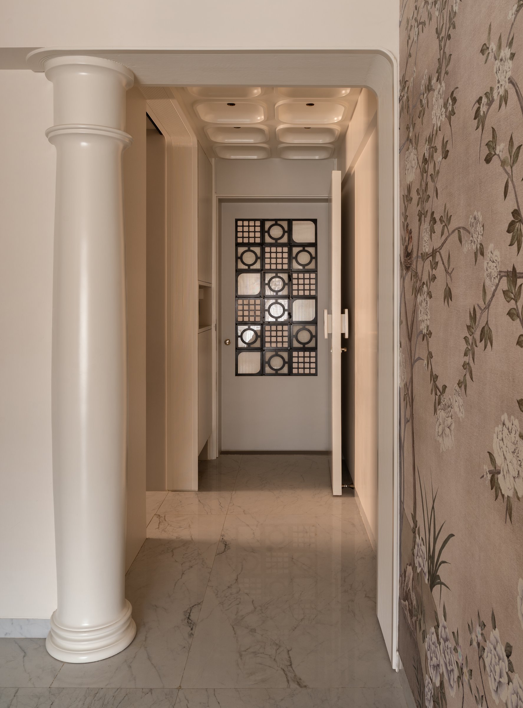 The foyer, where a freestanding column narrows the passage into the living room, framed by chinoiserie wallpaper and a grille-fronted door beyond