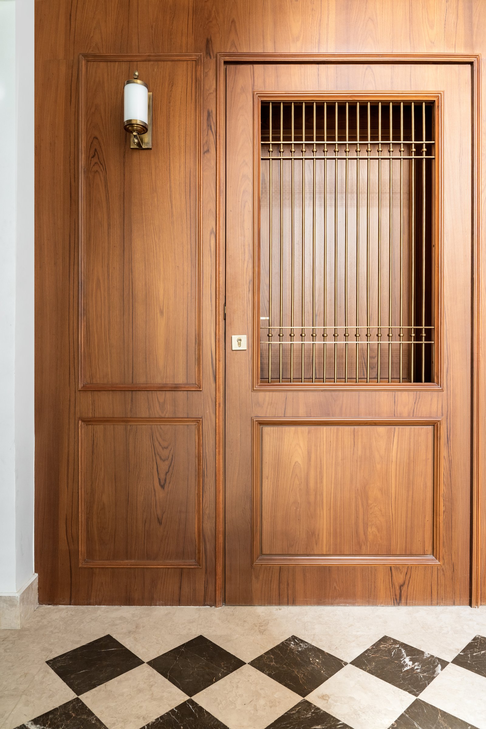 The entry, where a panelled teak door with a slender brass grille sits above a chequerboard floor of travertine and dark marble