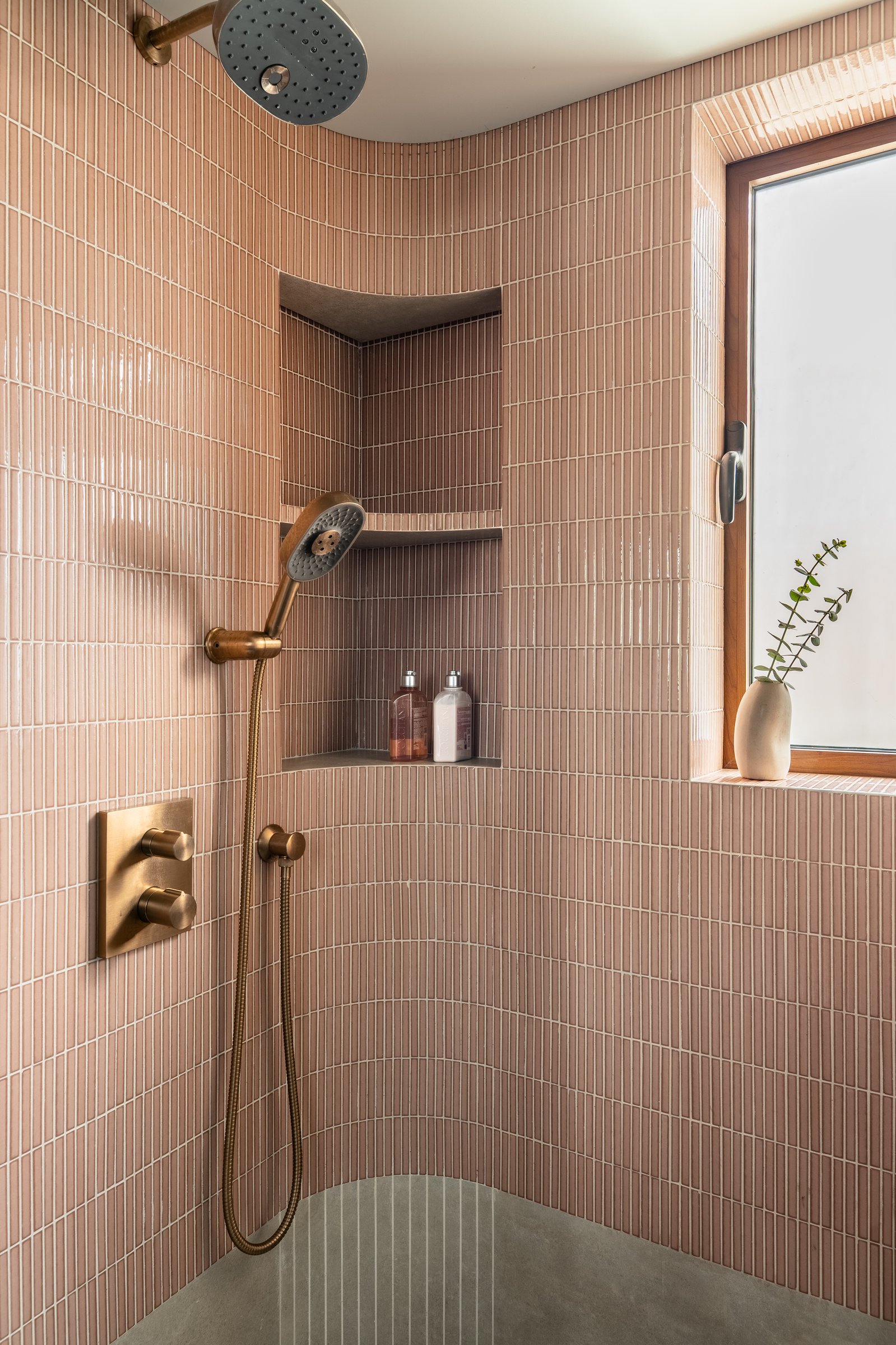 Within the shower, aged brass fittings warm the pink tiled field beside a timber-framed window