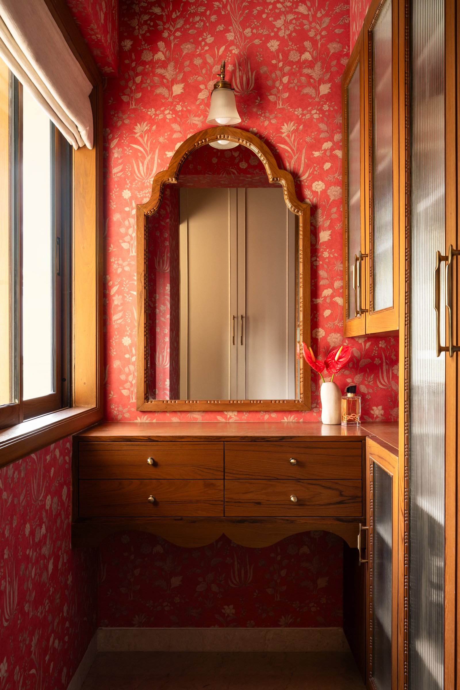 A dressing corner wrapped in red botanical wallpaper, with a carved arched mirror above a four-drawer floating vanity