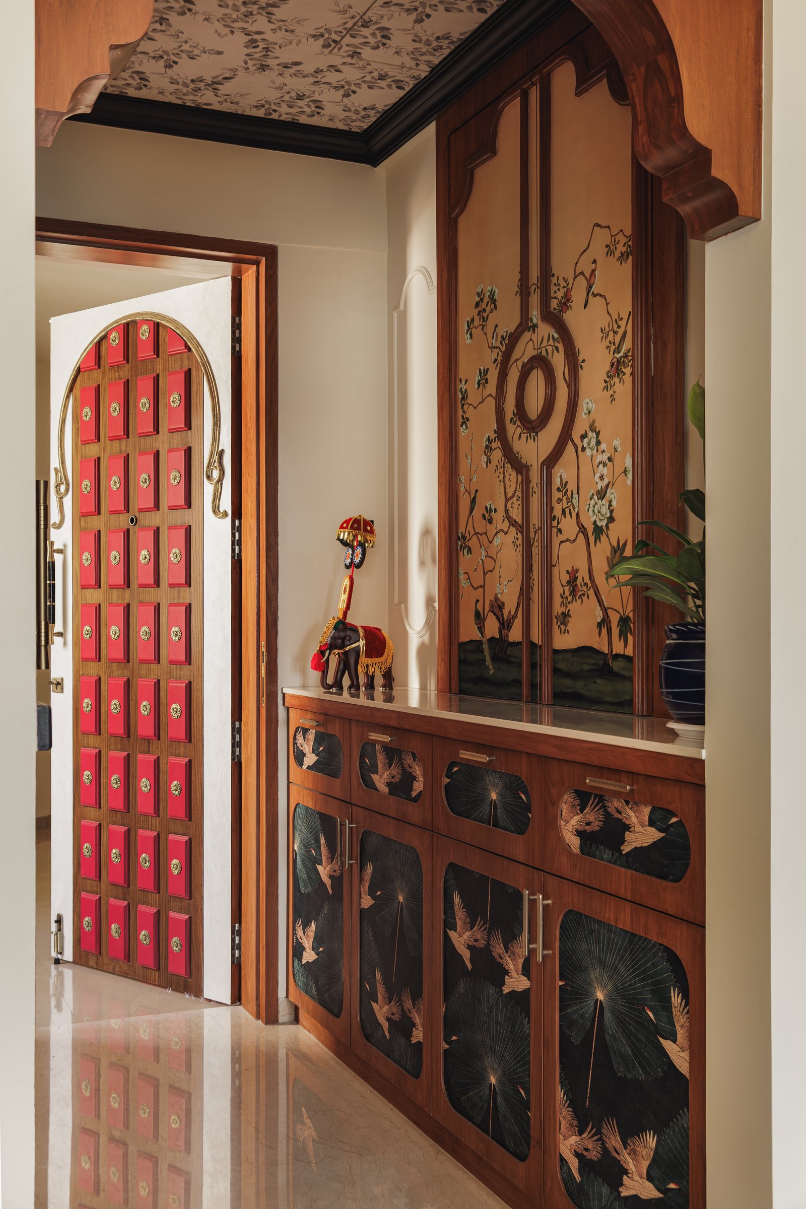 The foyer, where a crimson brass-studded door and a chinoiserie shoe console establish the home's layered material language