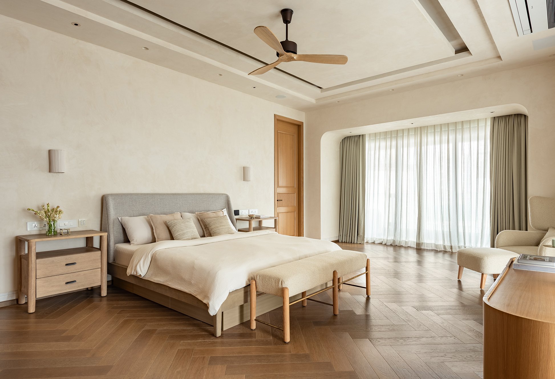 The bedroom opens onto the sea through sheer-and-sage drapery, with herringbone oak flooring extending the warmth of the plaster walls