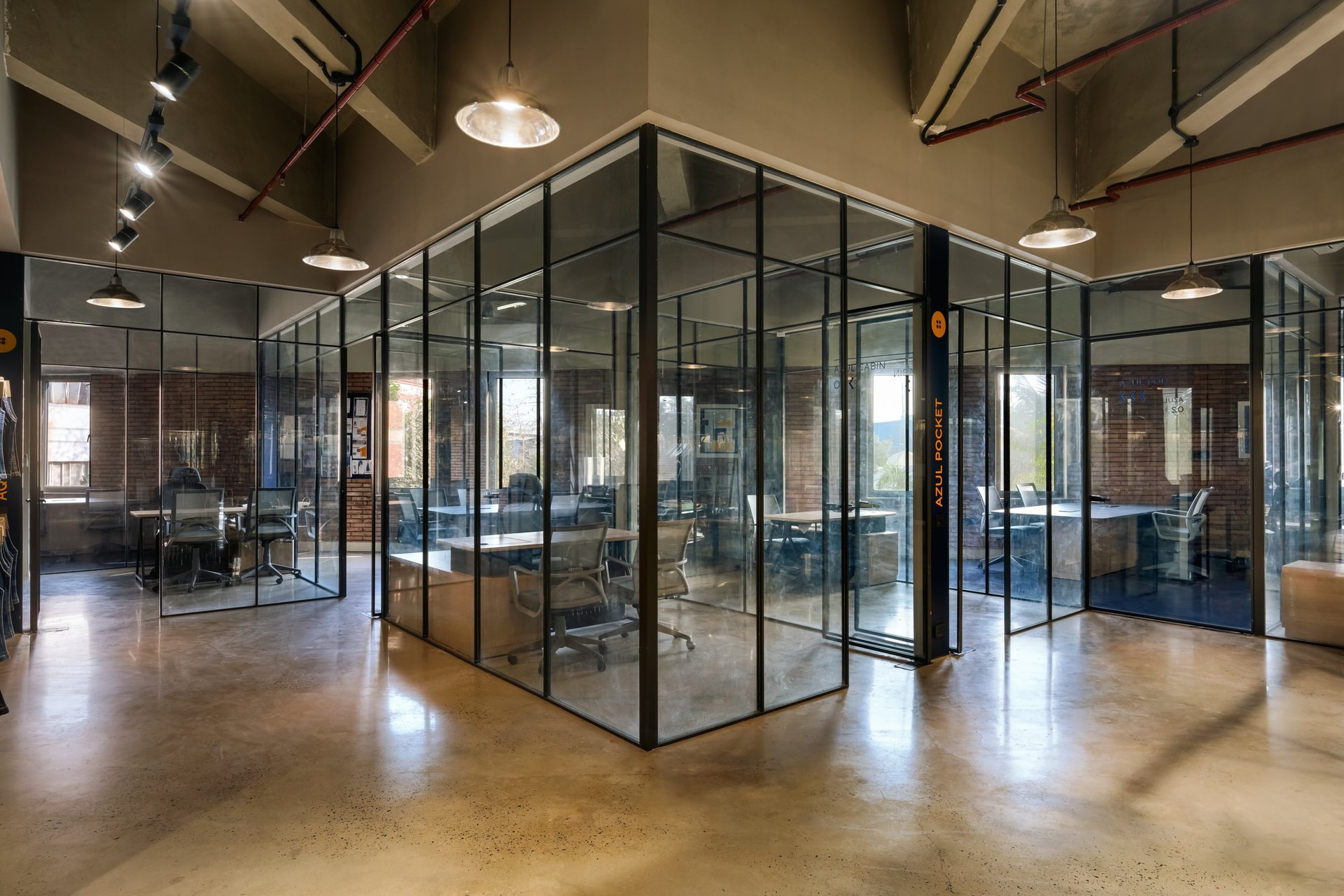 The peripheral office cluster, where steel-framed glass cabins ring an open circulation zone beneath the retained industrial ceiling