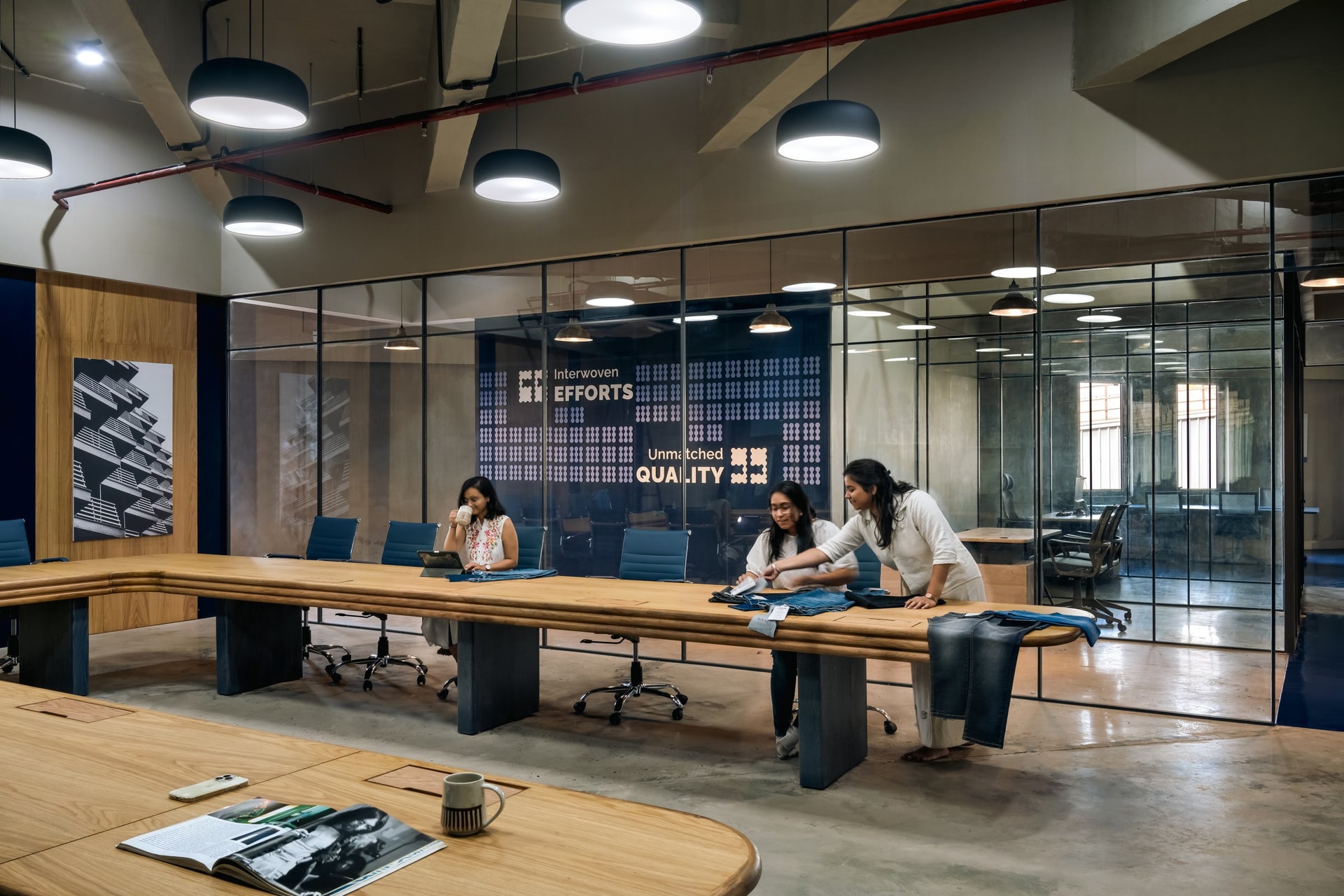 The boardroom, with its substantial U-shaped wooden table set beneath oversized pendants and framed by the 'Interwoven Efforts, Unmatched Quality' graphic
