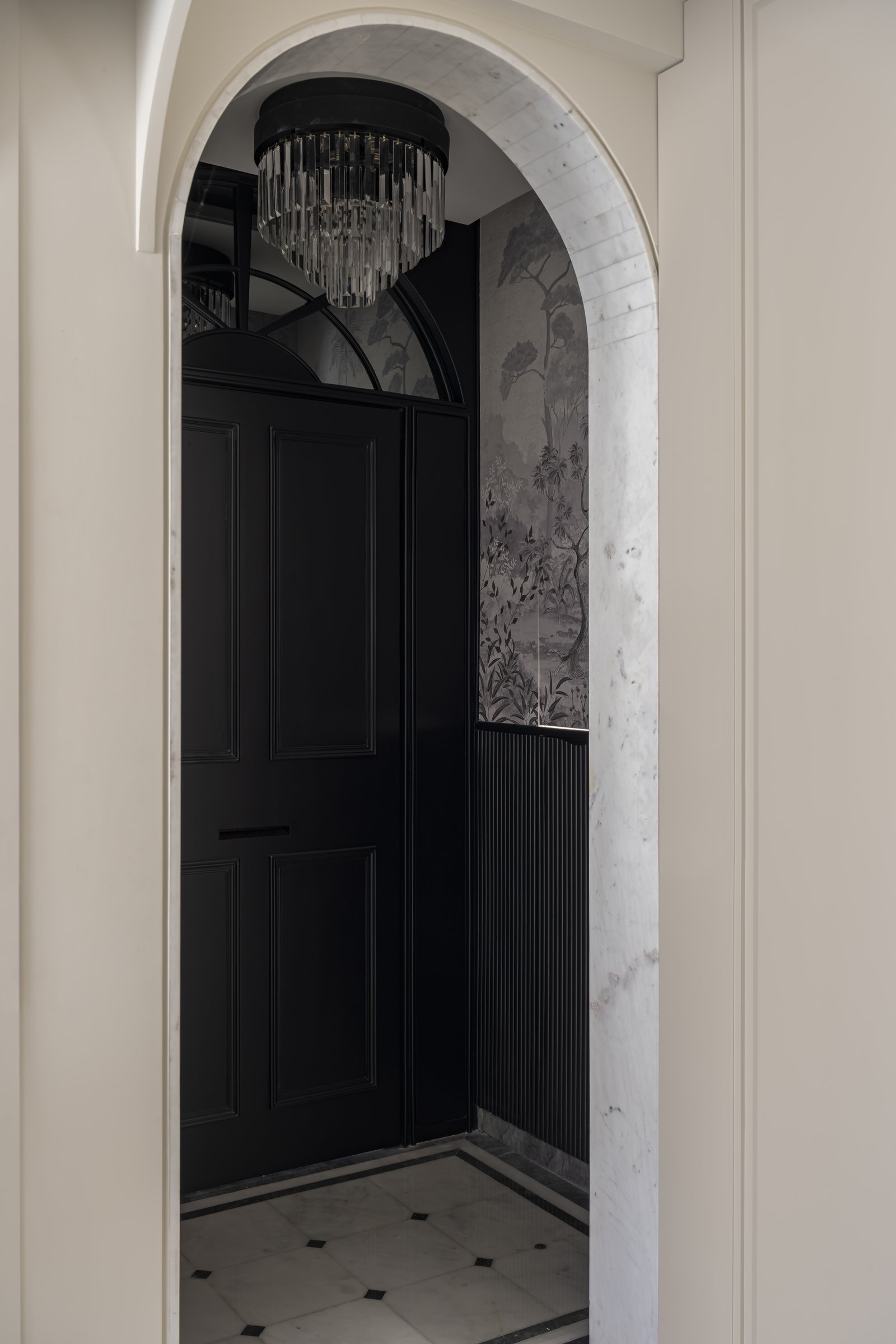 The entry foyer framed through its arched marble surround, with a panelled London-style door, fluted dark panelling and a grisaille wallpaper from Kalakaari Haath