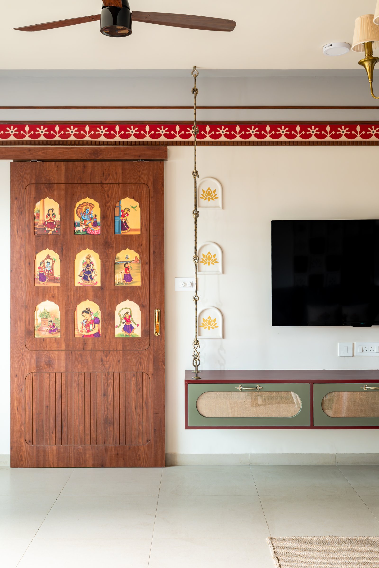 The media wall, where a sliding wooden door with painted figurative panels conceals the puja room beside a floating console in deep red, sage and cane