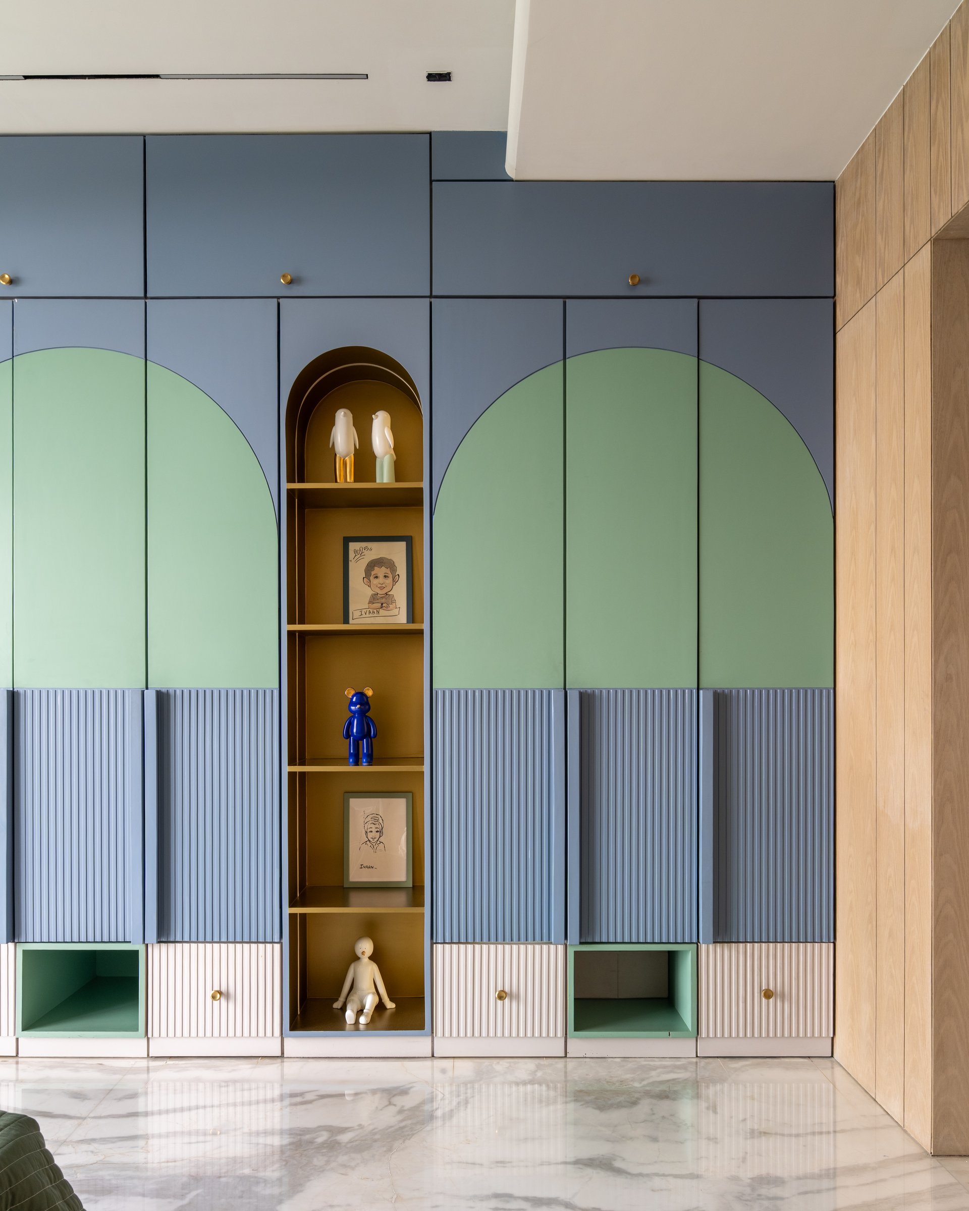The wardrobe wall in the children's bedroom, composed of sage shutters, periwinkle fluted panels and a central ochre display niche