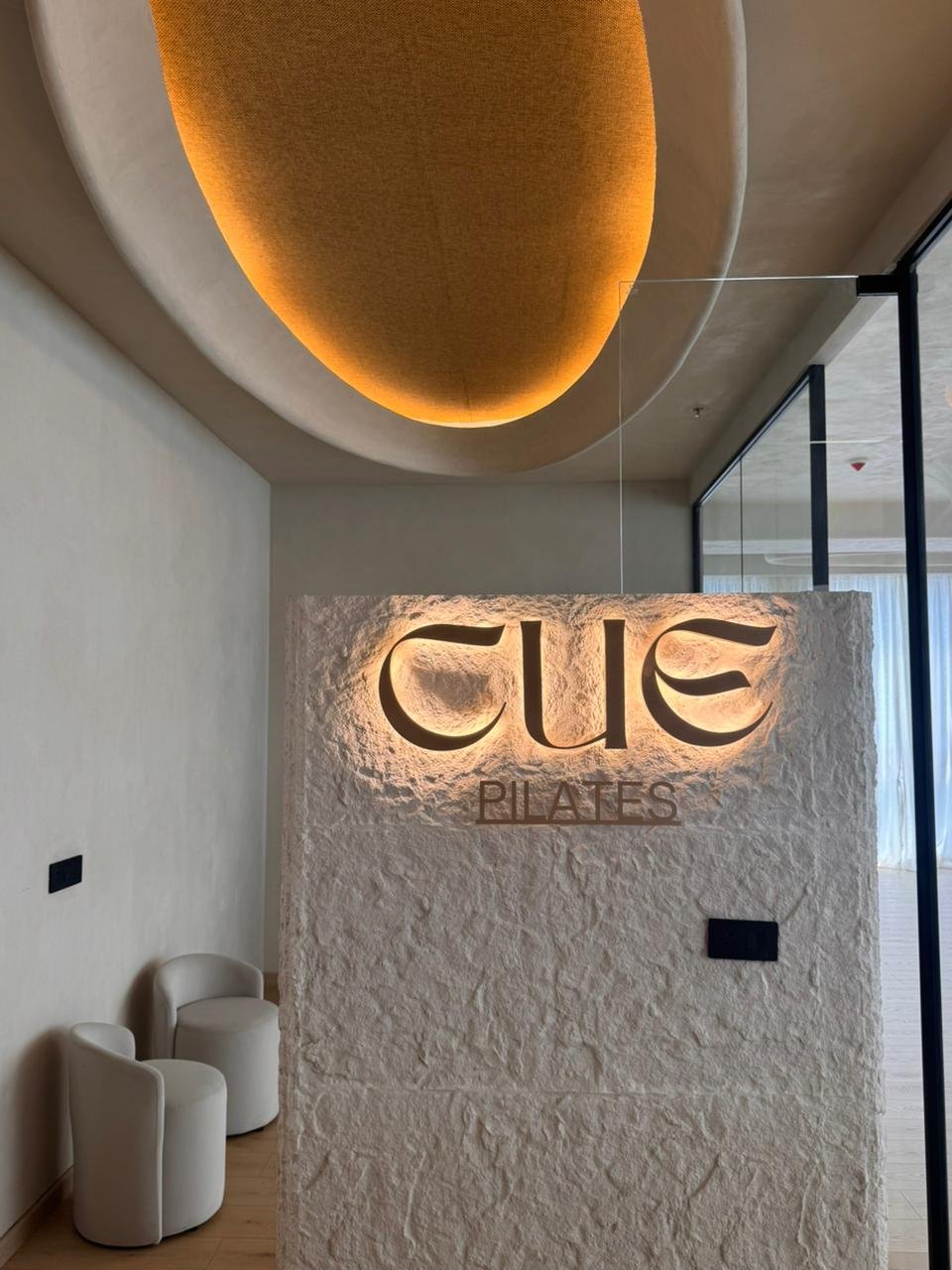 The arrival: a hand-textured plaster wall carries the backlit Cue Pilates wordmark beneath a softly glowing oval ceiling cove