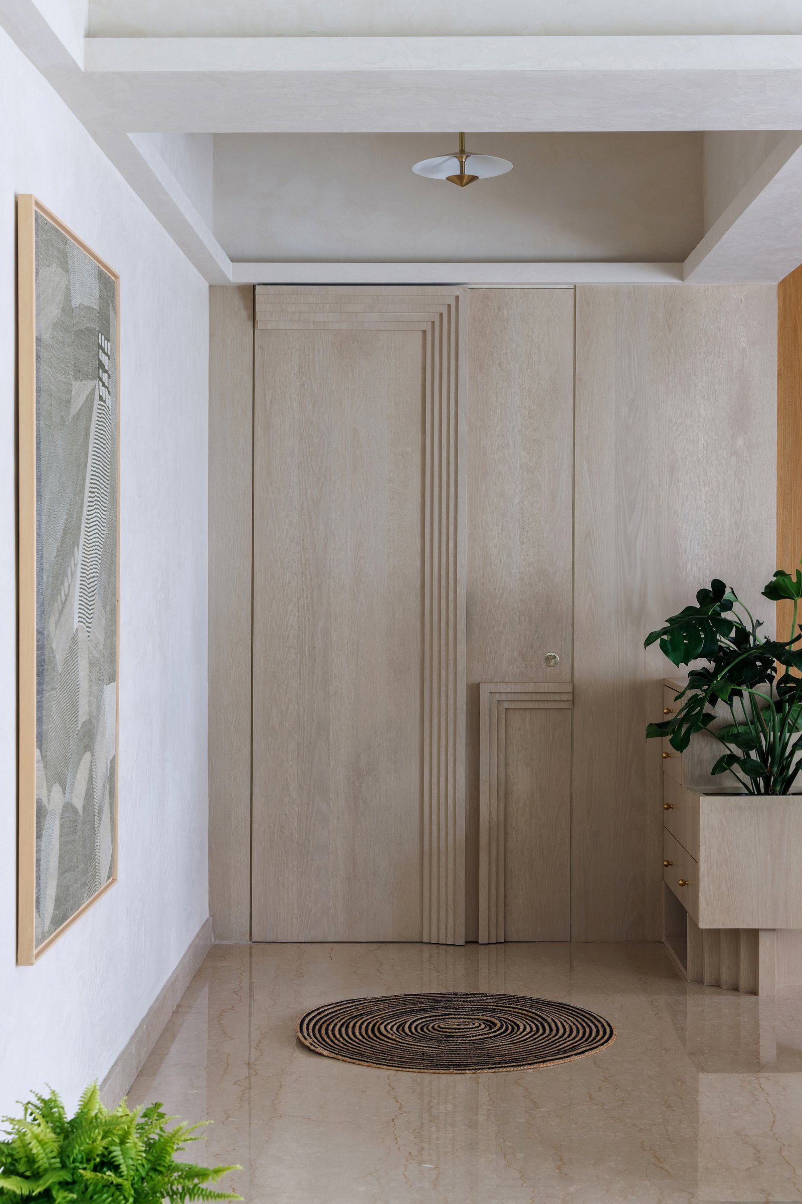 The foyer: a single pale ash plane absorbs the main door, shoe storage, and a concealed pocket entry, each opening articulated by a stepped, fluted reveal