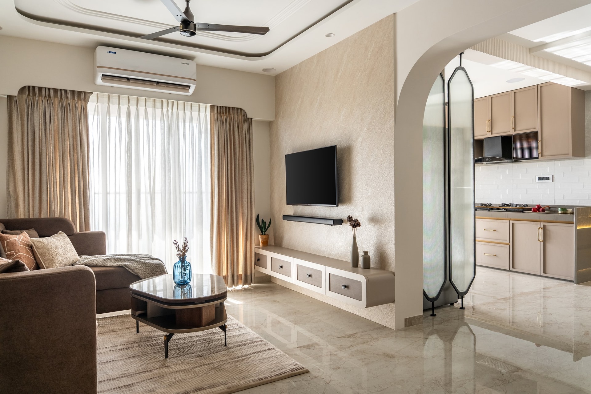 The living room opens through an arched glass partition into the kitchen, light sliding across marble and warming the cocoa upholstery